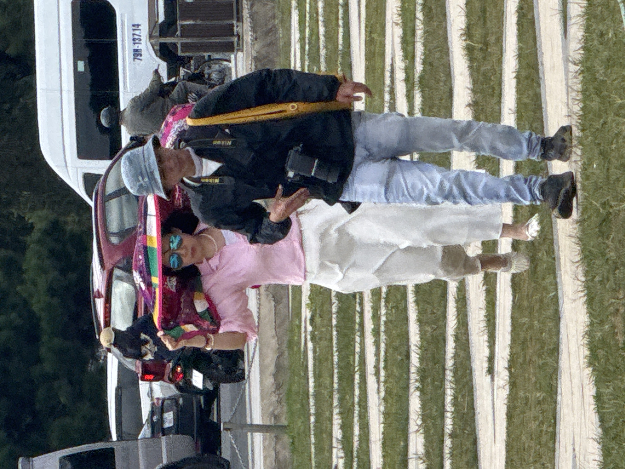In Da Lat, Vietnam, two individuals are walking on a paved pathway lined with grass. The person on the left, a woman, wears a pink long-sleeved shirt, white pants, and a wide-brimmed hat. She is holding a colorful, circular fan above her head with her right hand. She also wears sunglasses and a pearl necklace. The person on the right, a man, wears a blue bucket hat, a black jacket over a white t-shirt, and light blue jeans. A camera is attached to his jacket with a black strap, and a yellow strap crosses his shoulder. He is carrying a camera bag on his right shoulder. Behind them, a red SUV is parked, and a white van with the license plate "79H-137.14" is visible. A person on a motorcycle is in the background near the van. The environment appears to be outdoors, possibly a park or tourist area.
