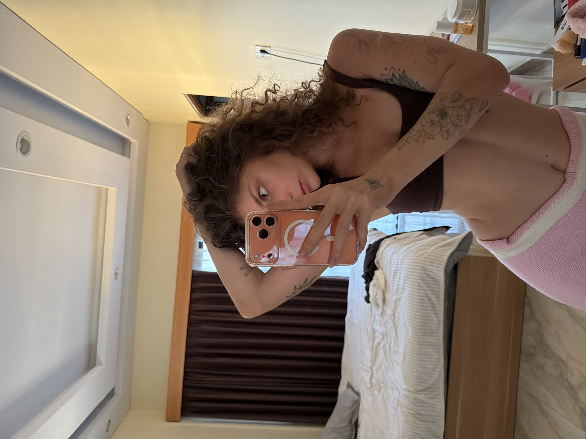 A woman is taking a selfie in a room. She has curly hair and tattoos on her arms. She is wearing a brown top and pink bottoms. She is holding a peach-colored phone. There is a bed behind her and a window with blinds. The room has a white wall on the left side. The location could be North Kuta, Indonesia.