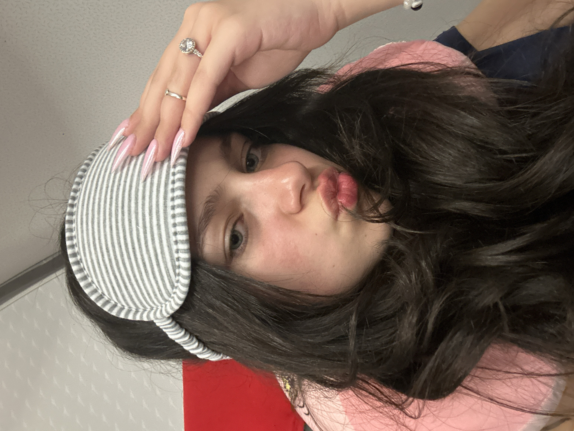 A young woman is posing for a selfie. She is wearing a striped eye mask on her forehead. Her long, dark hair is styled in waves. She is wearing two rings on her left hand, one with a large stone. Her fingernails are long and pink. She is pouting her lips. She is wearing a pink top. The background is out of focus but appears to be an interior space with a window. It is not possible to determine the location is Yerevan, Armenia from the image provided.