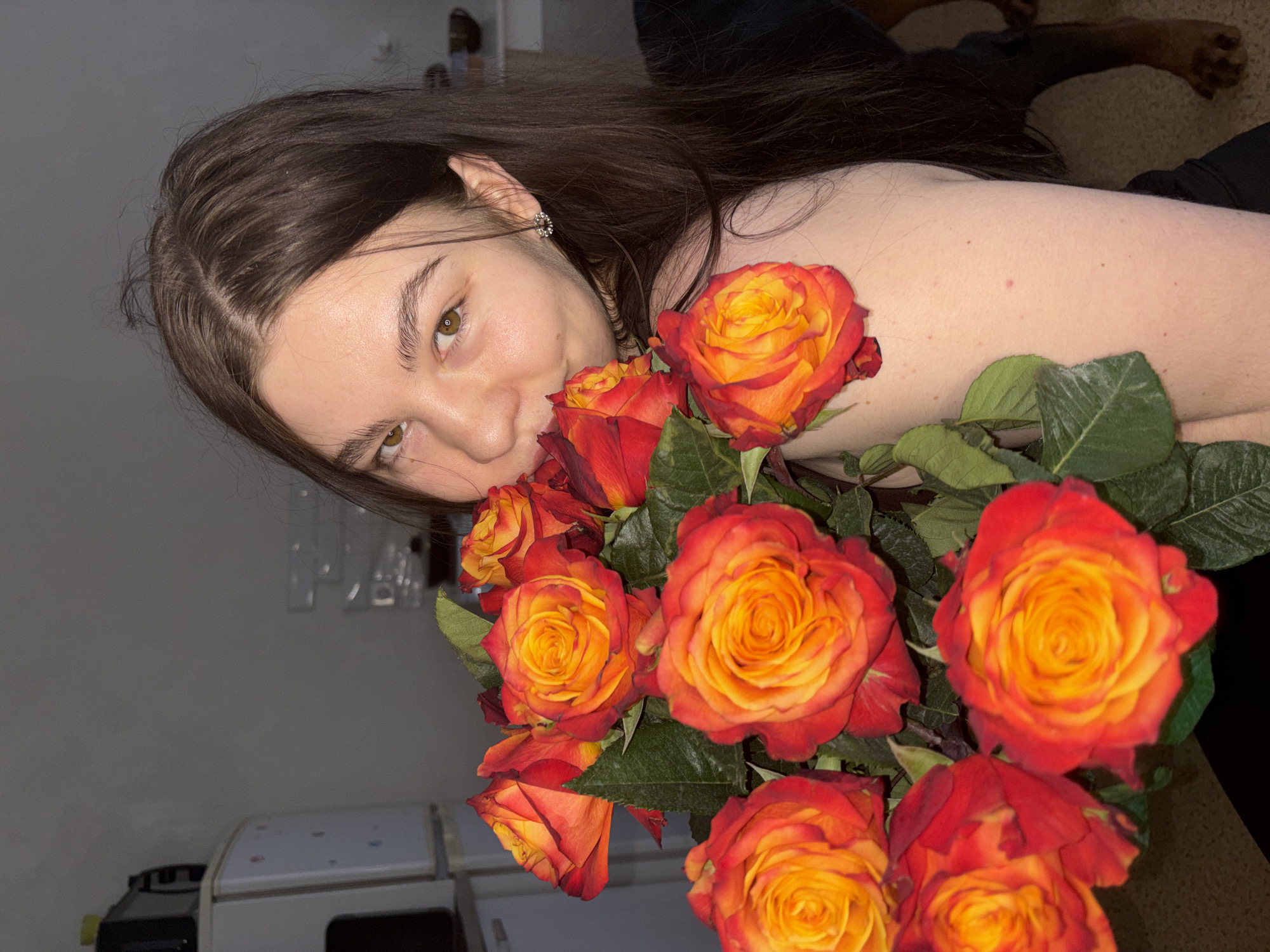 An angled indoor portrait features a young woman lying down, holding a large, vibrant bouquet of roses close to her face and chest. The woman, positioned in the lower left and center of the frame, has long, dark brown hair that cascades around her. She looks towards the camera with a gentle expression and a slight smile, her light brown eyes open. A small, sparkling earring adorns her visible left ear.
The dominant object in the image is the bouquet of roses, characterized by their striking bi-color petals that transition from a bright yellow-orange in the center to a rich reddish-orange or dark red at the edges. The roses are interspersed with deep green leaves. The woman's pose suggests she is either smelling, holding, or presenting the flowers. She appears to be wearing a dark, possibly strapless garment, with her shoulder and part of her chest visible.
The background is dimly lit and largely out of focus. A plain, light gray wall occupies the left side of the frame, with a hint of a light-colored appliance or furniture item in the bottom left corner. The upper right background shows a dark, textured surface, possibly carpet or clothing, with a blurry hand or arm visible. The overall lighting suggests an indoor setting, likely in the evening or at night. The scene conveys a sense of contentment or appreciation. Based on the provided context, the image location is Perm, Russia. No visible text is present in the media.