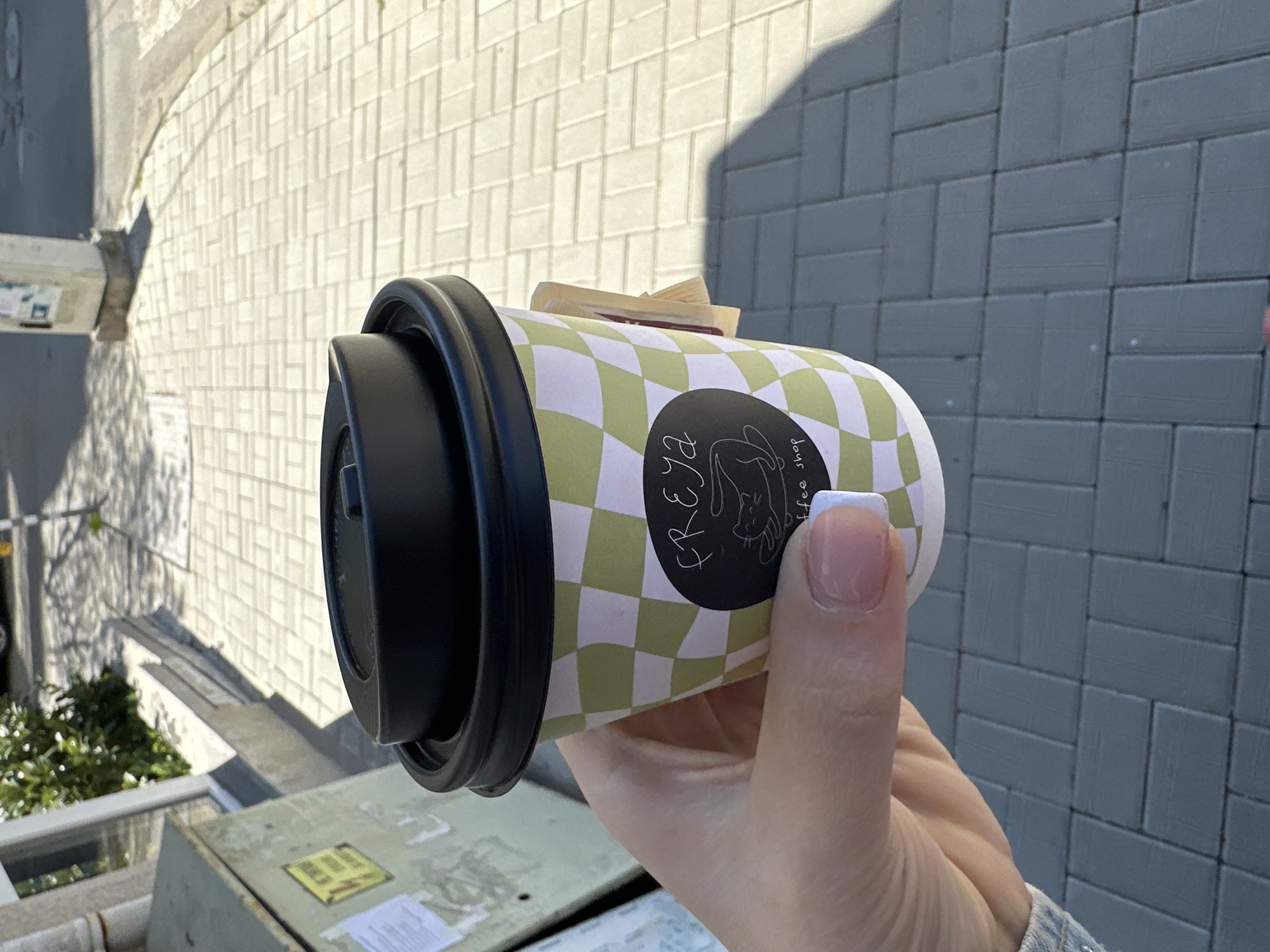 A hand holds a disposable coffee cup with a black lid and a green and white checkered pattern. The cup has a black oval logo with the name "FREYA" written vertically and a drawing of a rabbit inside. A folded paper napkin or item is sticking out of the top of the cup. The hand has manicured nails with white tips. The background consists of gray brick pavers and a view of the city, including a building and vegetation. The location appears to be a balcony or elevated area in an urban setting, possibly Sofia, Bulgaria, given the architectural style. A metal box or utility unit is visible below, near a window or opening with metal bars.