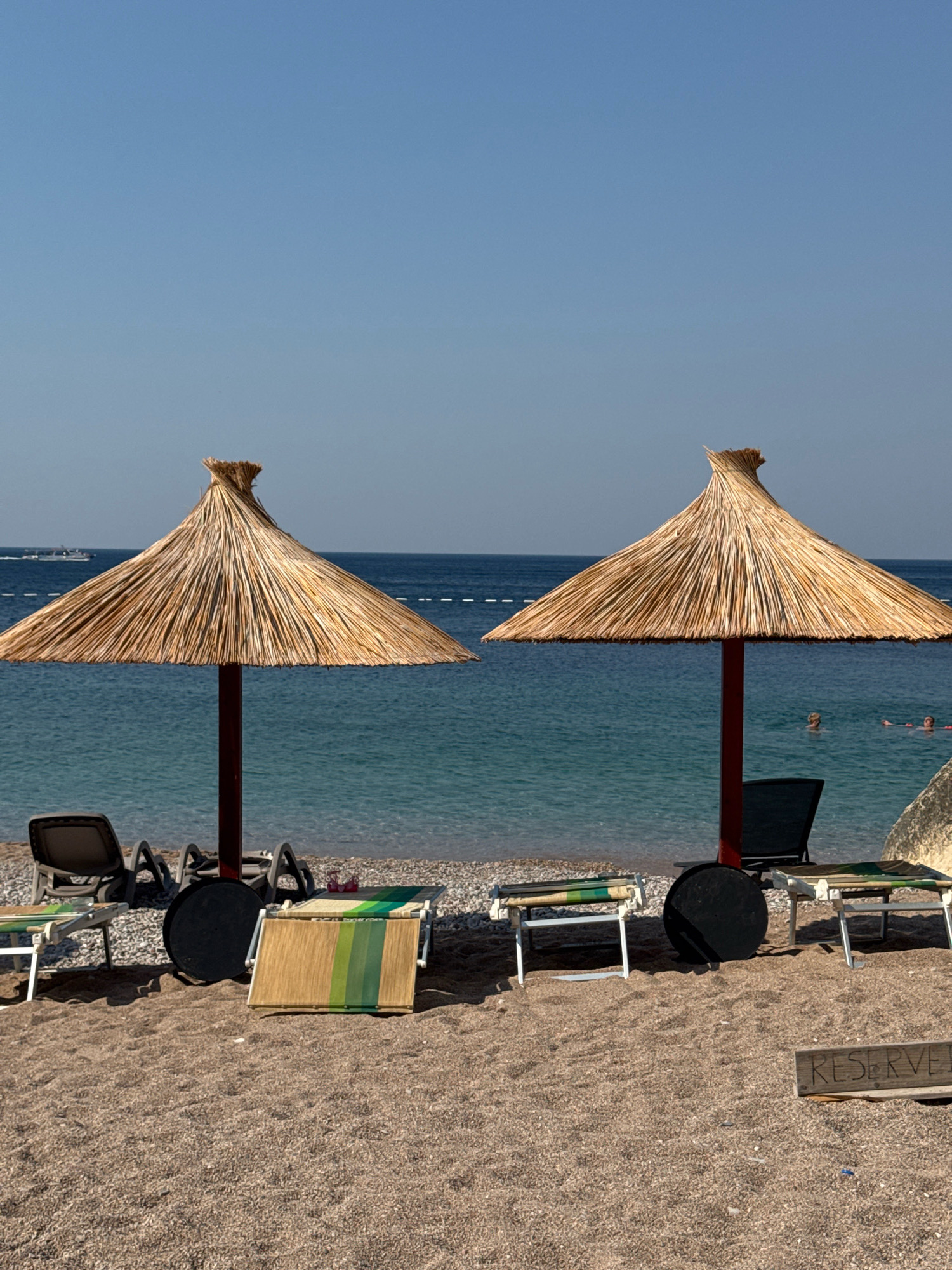 Two thatched umbrellas stand on a pebble beach in Mirište, Montenegro, with the Adriatic Sea in the background. The umbrellas provide shade for lounge chairs. Several lounge chairs are visible on the beach, some unoccupied and some with beach mats. In the water, a few people can be seen swimming. A boat is visible in the distance. A wooden sign on the beach reads "RESERVED". The sky is clear and blue.