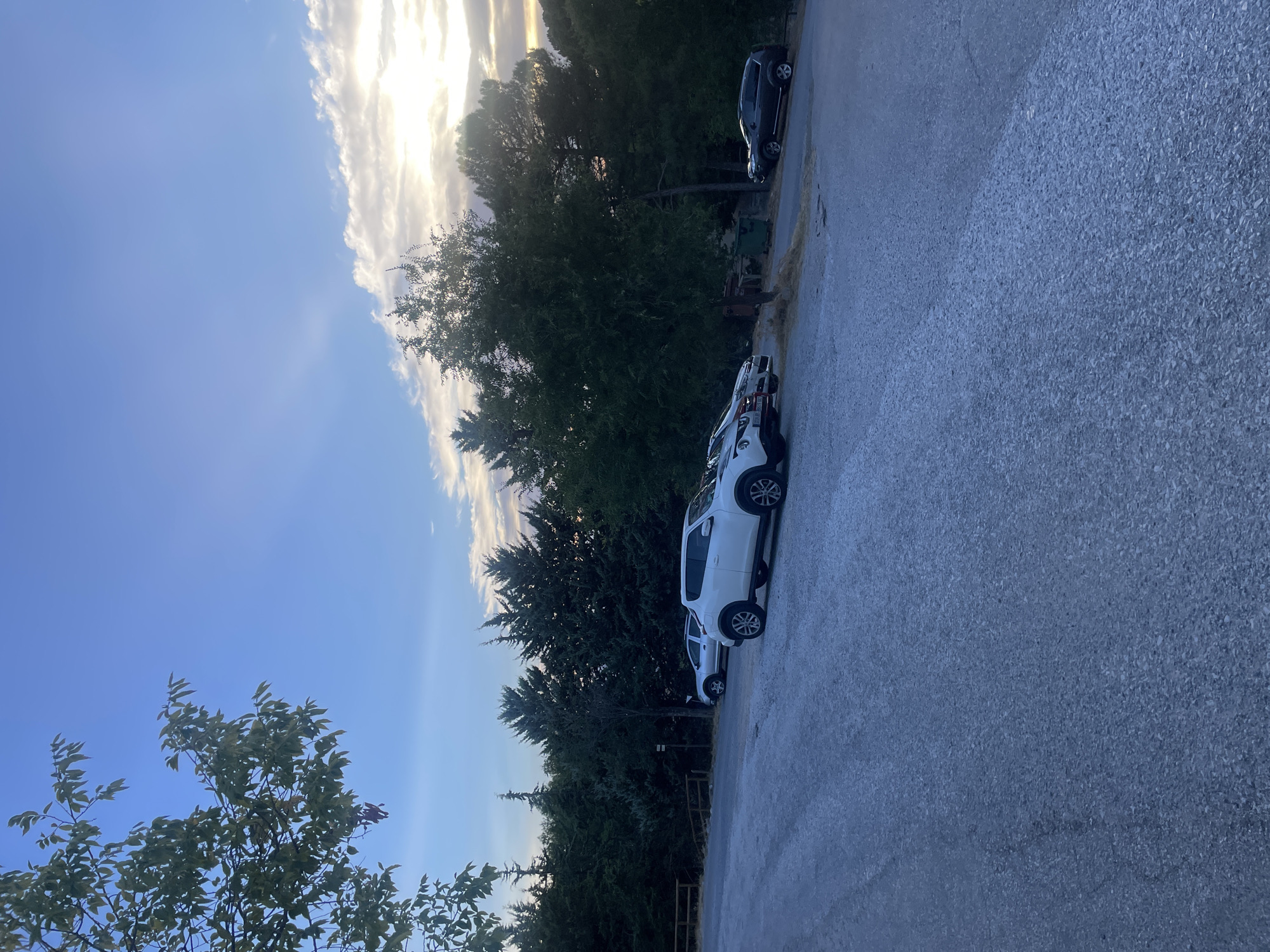 The image depicts an outdoor scene at a parking area situated in the city of Víznar, Spain. The time of day appears to be late afternoon or early evening, as indicated by the soft, diffused sunlight casting through a partly cloudy sky. The foreground is dominated by a gravel and asphalt surface, indicative of a parking lot.
Several vehicles are parked in the lot. A white compact SUV is prominent in the center-right of the frame, facing to the left. To its left, a silver sedan is parked. Further to the right and slightly further back, a dark-colored SUV is parked, also facing left. The trees lining the back of the parking lot are lush and green, with a dense canopy creating a shaded backdrop. A wooden fence is visible to the far left, bordering the parking area. The sky is a gradient of blue, transitioning to lighter tones near the horizon where the sun is partially obscured by clouds, creating a soft, atmospheric light. There are no people visible in the image. The scene is tranquil and appears to be a common area for parking, possibly near a park or a natural reserve given the surrounding foliage.