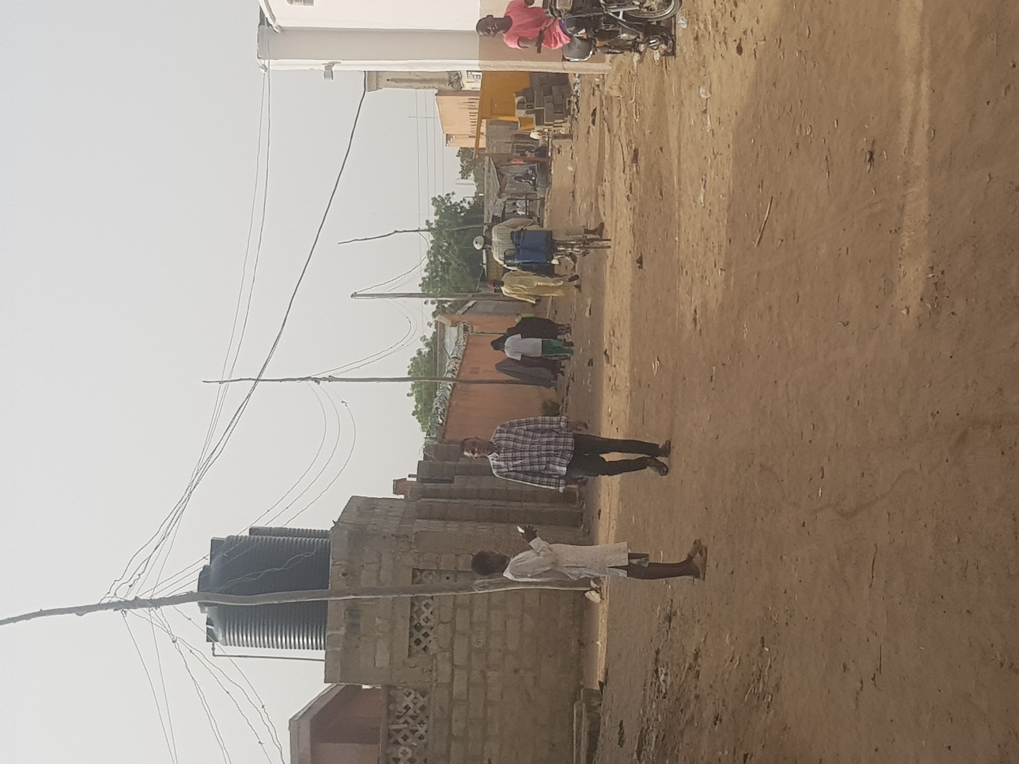 The image depicts a street scene in Dabawa, Nigeria. The ground is a dirt track, likely a road. In the foreground, a young person dressed in white stands. Further along the road is a man wearing a plaid shirt and dark trousers walking. Still further along, several people stand in front of buildings. A man on a bicycle rides on the street, and another man on a motorbike drives towards the camera. Buildings line both sides of the road, interspersed with trees. Utility poles and wires stretch across the sky. A black water tank sits on a building to the left.