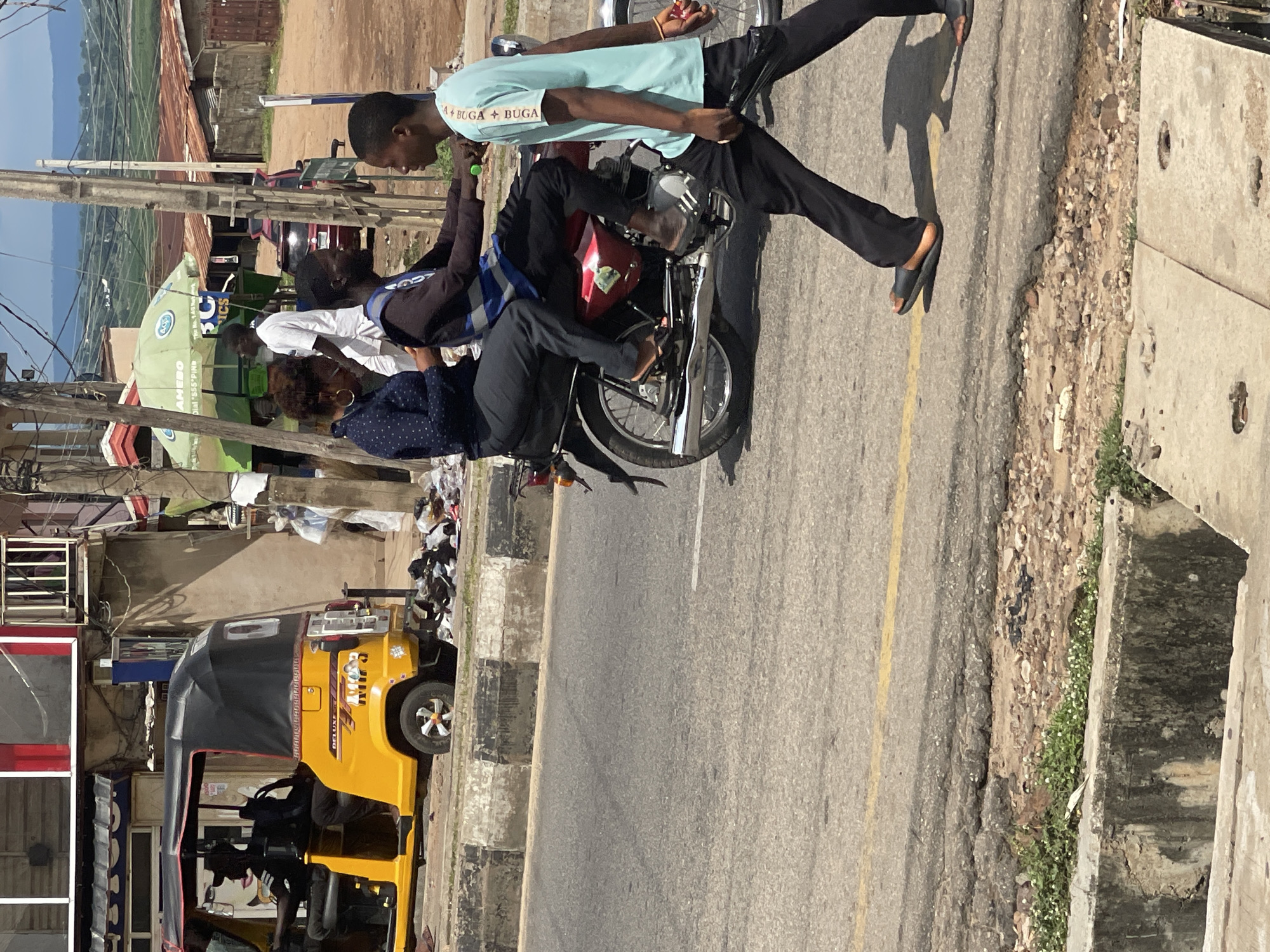 In an outdoor urban environment, a motorcyclist wearing a blue and black vest and black pants drives a red motorcycle. A female passenger in a blue patterned shirt and black pants sits behind the driver. A young male in a light blue t-shirt and black pants walks in front of the motorcycle, looking to his left. A yellow auto-rickshaw with a black canopy is parked to the left, with a person in a black cap driving. The background shows buildings, including shops with awnings, and a green umbrella over a vendor stall. Power lines are visible overhead, and a hillside with scattered structures and vegetation is in the distance. The ground is asphalt with a yellow line, and there is a concrete curb and a drainage ditch. Piles of trash are visible near the auto-rickshaw.