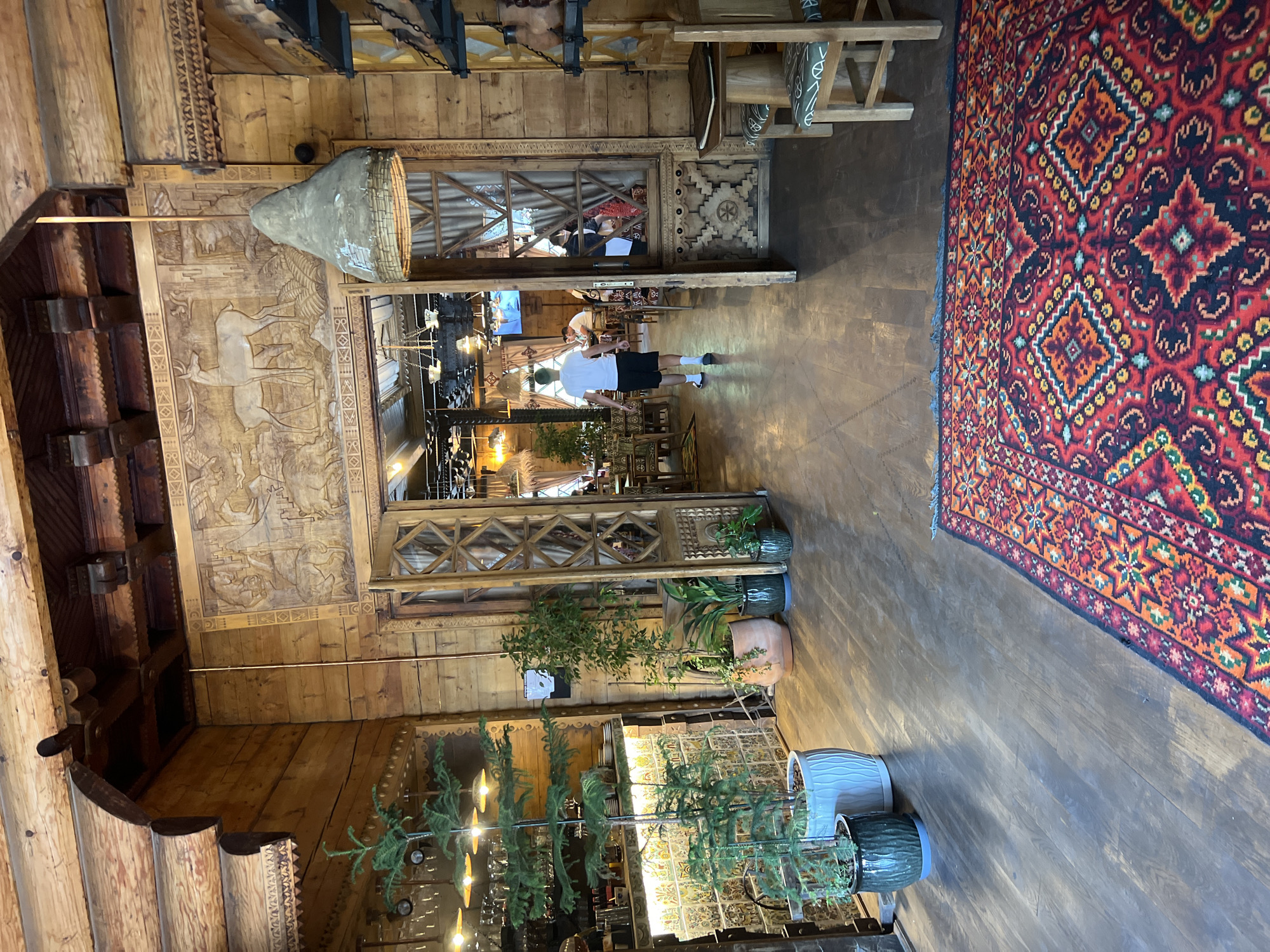 The image shows the interior of a rustic wooden restaurant in Yaremche, Ukraine. The walls, ceiling, and entryway are constructed from natural wood, with intricate carvings and decorative elements. In the foreground, a large, vibrantly colored Persian rug with geometric patterns covers the wooden floor. Several potted plants are strategically placed throughout the space, adding touches of greenery.
The main entryway, framed by carved wooden doors, leads into another section of the restaurant. Through the open doors, several tables and chairs are visible, as well as patrons dining and conversing. A man in a white t-shirt and black shorts is walking away from the camera into the restaurant. A distinctive lampshade, made of what appears to be rough plaster or clay with a woven detail at the bottom, hangs from the ceiling in the central part of the entryway.
To the left of the entryway, a bar area is partially visible, stocked with glassware and bottles. Decorative tiling adorns the wall behind the bar. The overall ambiance is warm and inviting, with a strong emphasis on natural materials and traditional design.