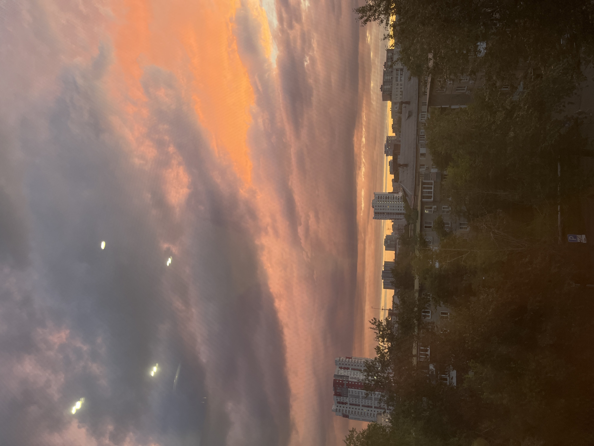 This image captures a dramatic sunset over a residential area in Chelyabinsk, Russia. The sky is filled with a riot of colors, from deep grays and purples in the foreground clouds to vibrant oranges and pinks higher up, suggesting the last rays of sunlight breaking through. The dramatic cloud formations create a sense of awe and a touch of melancholy.
Several tall apartment buildings are visible, their windows reflecting the fading light. Some buildings are modern, with colorful facades, while others appear older and more utilitarian. Dense trees line the foreground and are interspersed between the buildings, their leaves dark against the colorful sky, adding to the atmospheric depth.
The image appears to be taken from a slightly elevated position, possibly a balcony or window, looking out over the urban landscape. The lighting suggests it is late evening, nearing dusk. There are no people or specific activities clearly depicted, allowing the focus to remain on the natural spectacle of the sunset and the urban environment it illuminates. A small blue and white sign, possibly a parking sign, is visible on a pole near the top right of the image, providing a subtle urban detail. The overall mood is one of serene beauty mixed with the quietude of a city settling in for the night.