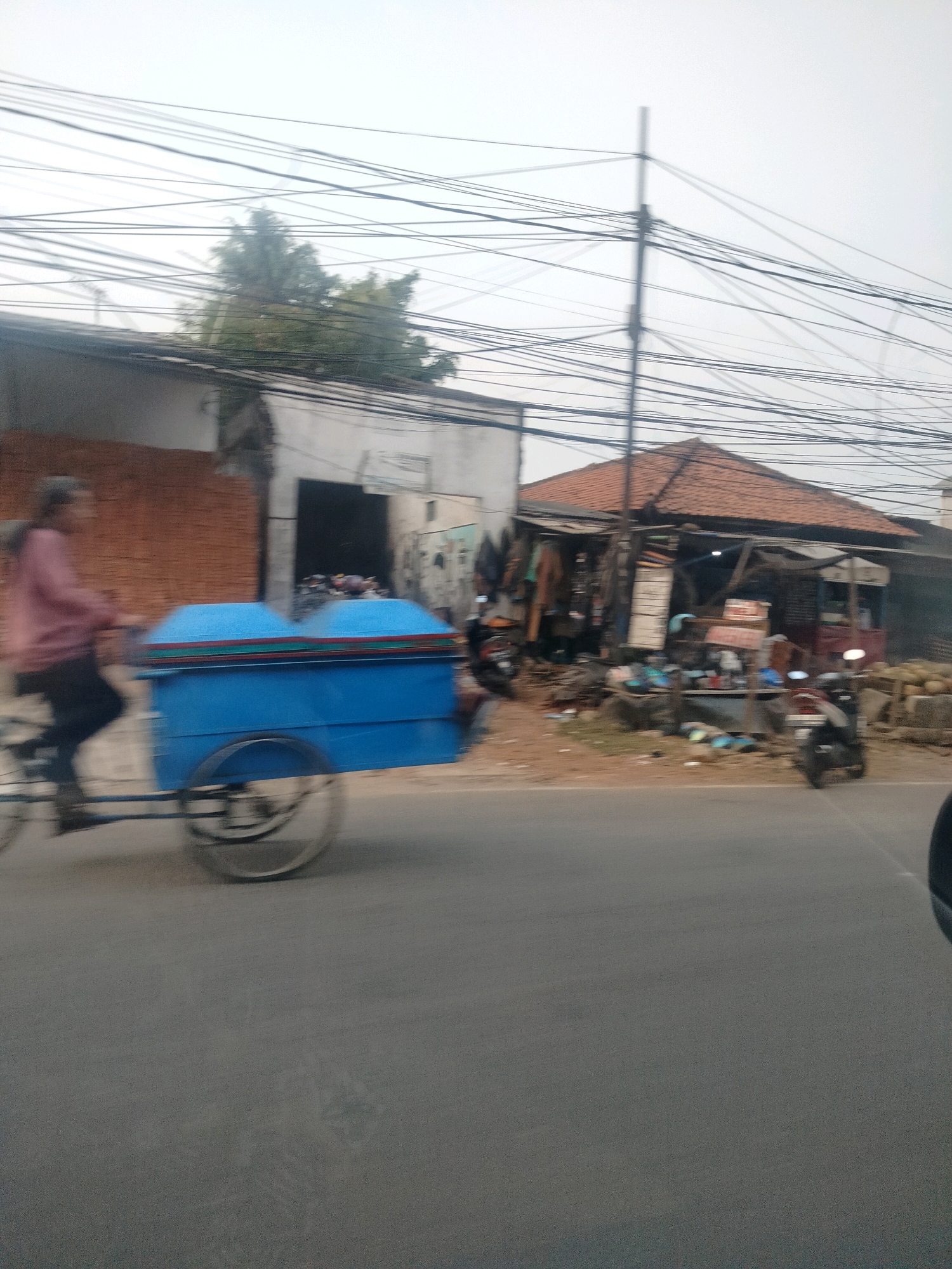 In East Jakarta, Indonesia, a person rides a bicycle pulling a blue cart with two rounded lids, possibly for food or goods. The cart is decorated with horizontal stripes of red, green, and yellow. The person is wearing a pink shirt and dark pants. In the background, a small building with a tiled roof and an open-air market area is visible. Several motorcycles are parked in front of the market. A dense network of electrical wires spans across the sky above the scene. The overall environment appears to be a street scene in a densely populated urban area, with elements of commerce and daily life. The image is slightly blurred, suggesting movement.