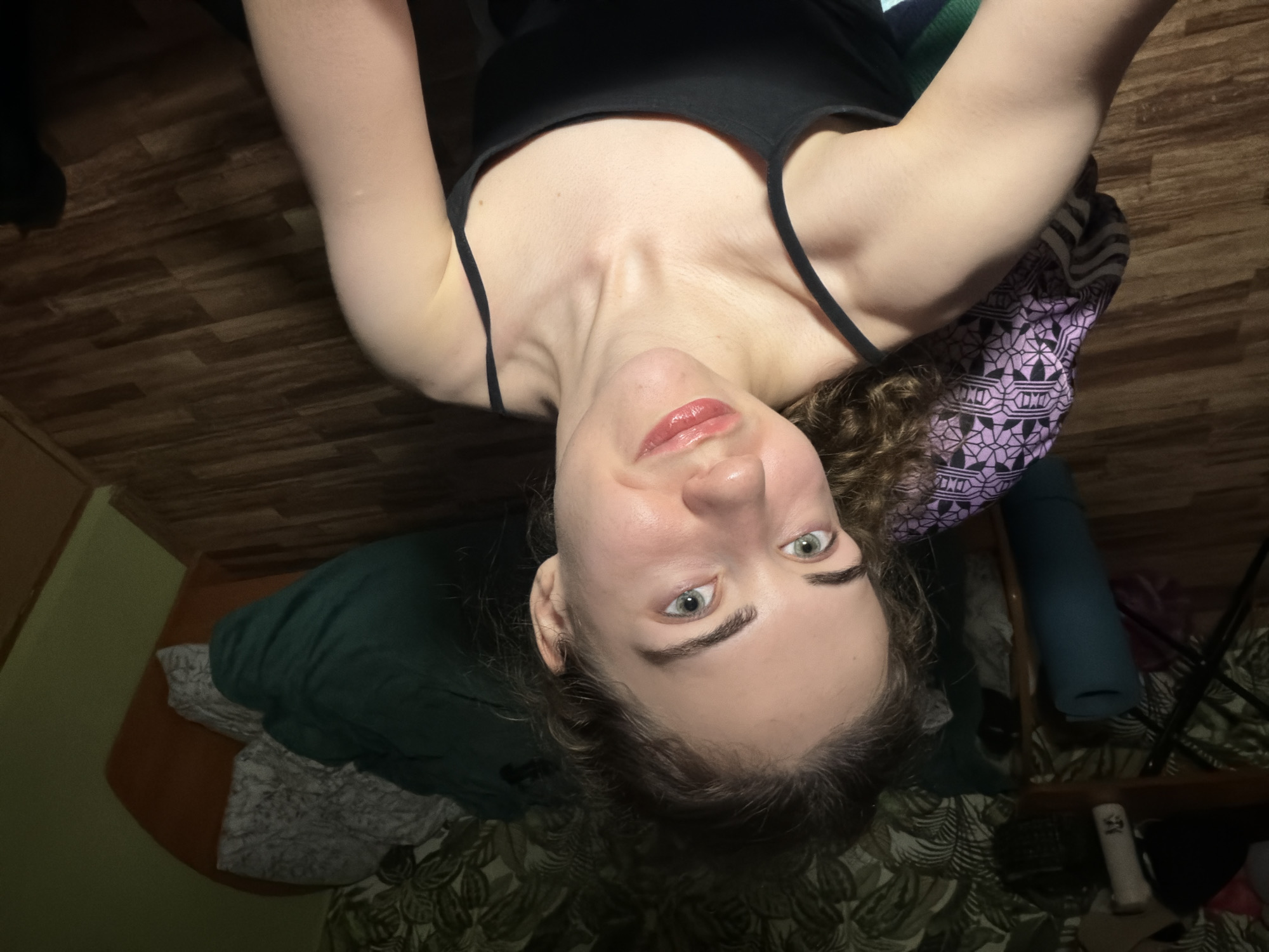 The image shows a young woman with light skin, light brown curly hair, and green eyes. She is wearing a black tank top and sitting in a chair with a patterned cushion. Her lips are slightly parted and appear to be glossed. The background suggests an indoor setting, possibly a bedroom, with a bed covered by a dark green blanket and a patterned duvet. There is a wooden floor and a wall with a leafy wallpaper. A yoga mat is visible in the left side of the frame, along with a table and some objects on it. The lighting is dim and appears to be artificial. The scene is located in Zavodoukovsk, Russia.
