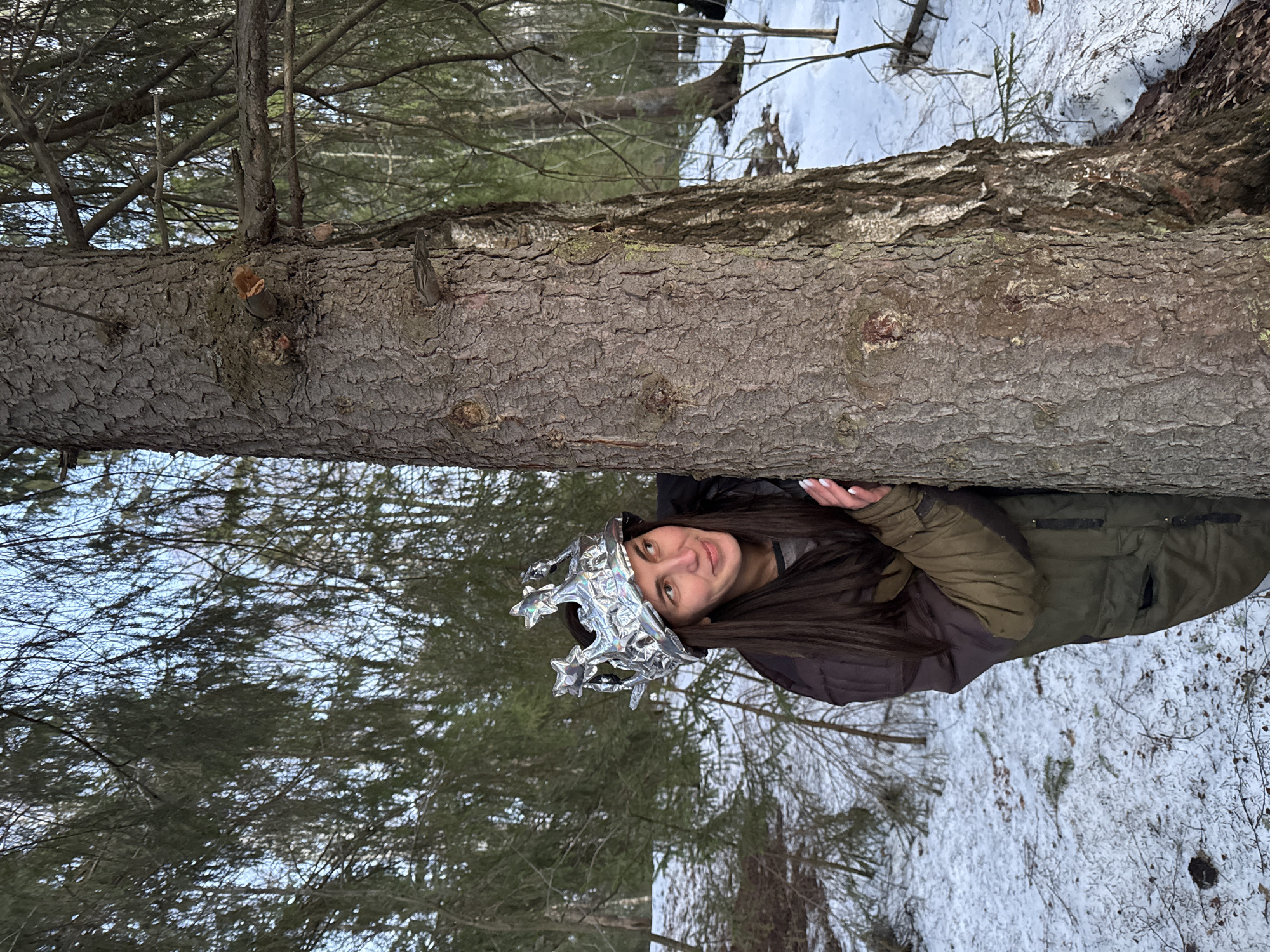 The image shows a woman in an outdoor location, likely in a forest or wooded area near the city of Вассята, Russia. There is snow on the ground. She is wearing a silver crown and is positioned behind a fallen tree trunk. The woman appears to be partially obscured by the trunk, with her head and torso visible. She is wearing a dark jacket with olive green sleeves. The background consists of trees and snow-covered ground.