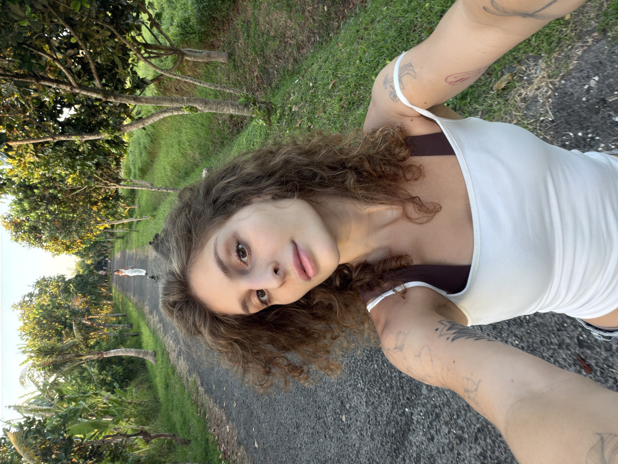 A medium-shot selfie shows a young woman with curly brown hair and light skin, wearing a white tank top over a dark sports bra. Her right arm, visible with tattoos, is extended towards the camera, suggesting she is holding the phone. She is standing on a narrow paved road. In the background, there are trees on both sides of the road, with lush green grass visible under them. A person in white clothing is visible in the distance, walking away on the road. The trees and foliage indicate a rural or semi-rural environment. There is no identifiable information that confirms the location as Kediri, Indonesia. The environment shows a road, trees, green grass.