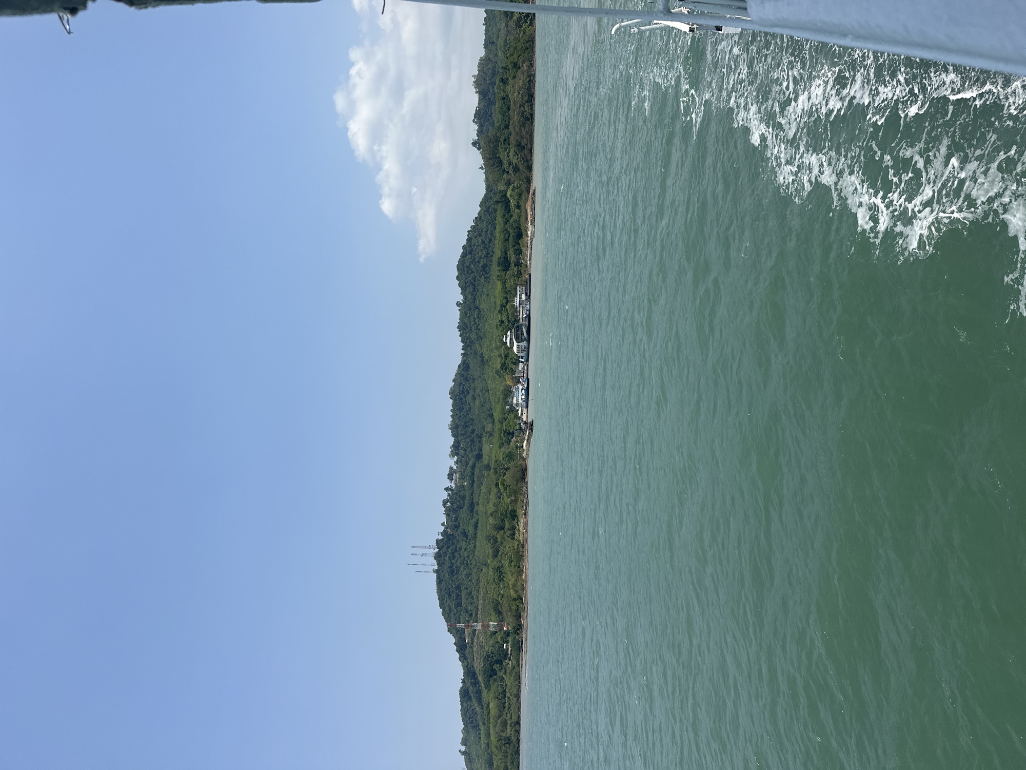 The image depicts a daytime scene in Laem Ngop District, Thailand, viewed from a boat. The sea occupies the right side of the frame, marked with white wake foam suggesting the vessel's movement. A coastline with dense green vegetation covers the left side of the frame. A few buildings are visible along the shore. There are a few antennas. The upper-left of the image shows a clear blue sky, punctuated by a white cloud. The top edge of the frame shows part of a metal railing, presumably of the boat the picture was taken on.