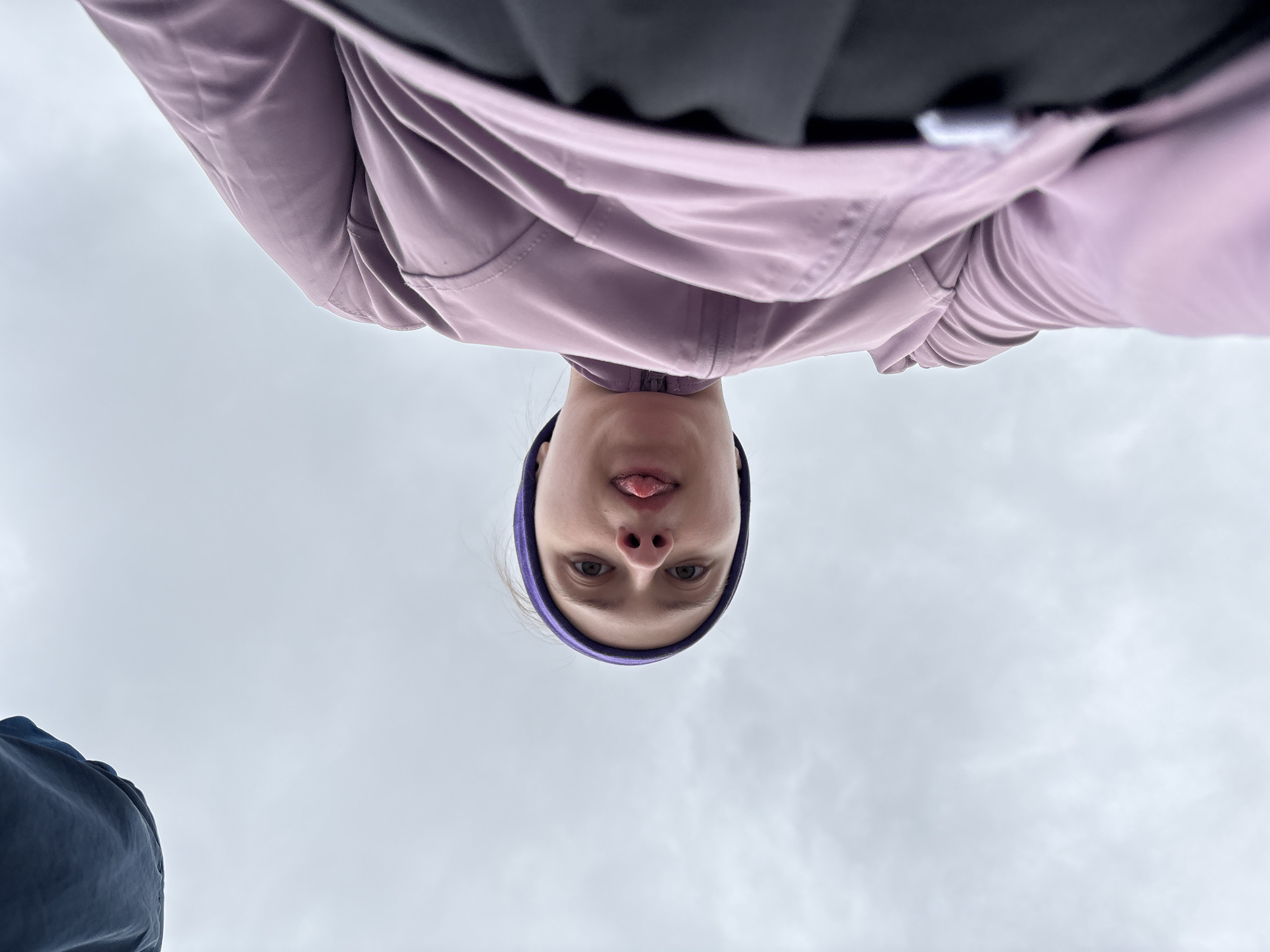 The image is an upside-down perspective shot. At the top of the frame is a person, likely female, wearing a lavender jacket and a purple headband. Part of a dark garment is visible behind the jacket. The person is sticking their tongue out slightly. Below them is a cloudy sky, and the corner of a blue jacket is visible on the lower left. The location is not identifiable as Perm, Russia from this image.