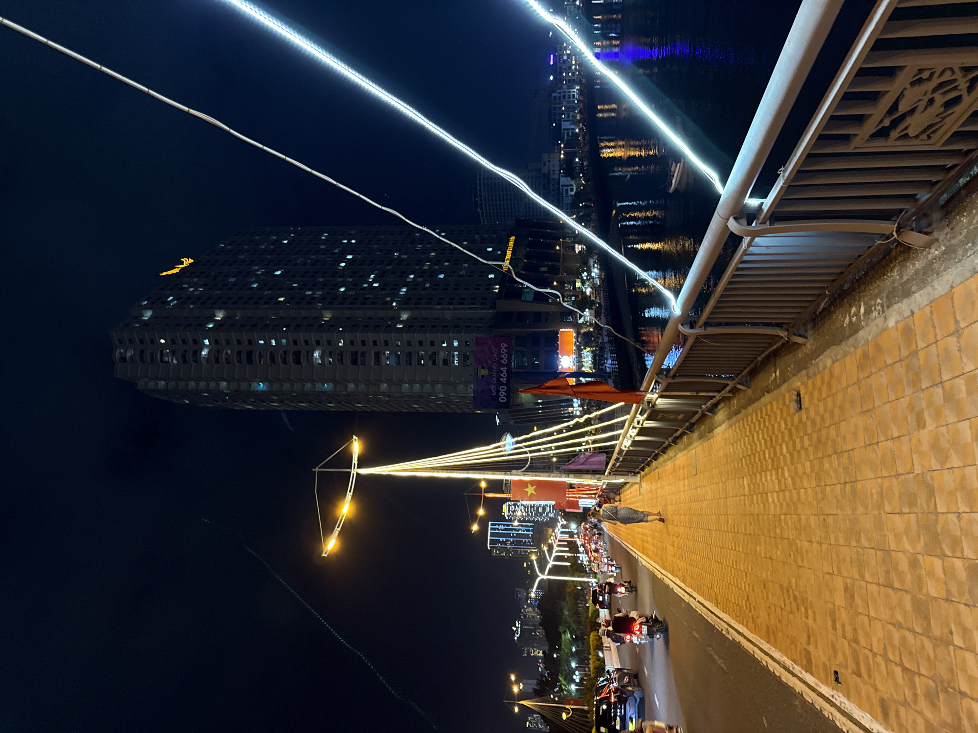 This is a nighttime, eye-level, medium-shot photograph of a pedestrian walkway on a bridge in Nha Trang, Vietnam. The walkway is paved with textured, golden-yellow bricks and bordered by a metal railing. On the left of the walkway is a street with traffic, including cars and motorcycles. The street is lit by streetlights and the headlights of vehicles. Beyond the street, the city skyline is visible, with numerous buildings illuminated by interior and exterior lights. In the foreground, a woman in a patterned dress walks away from the camera on the pedestrian walkway. A large, modern building dominates the background on the right, its numerous windows lit. The scene is illuminated by various light sources, including streetlights, building lights, and string lights that create streaks of light across the image. The water of a river or bay is visible to the right of the bridge, reflecting the city lights.