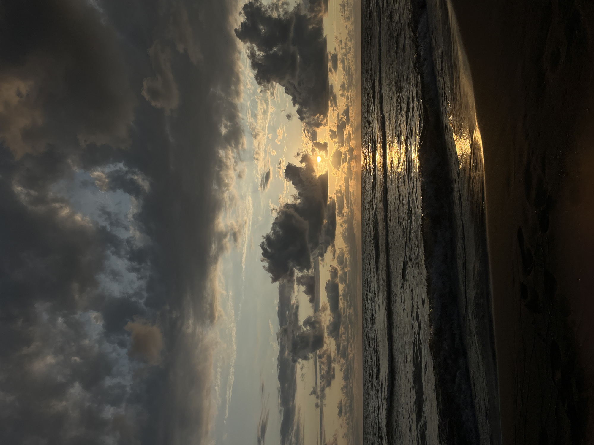 The image depicts a sunset over the Black Sea, viewed from a sandy beach in the village of Supsa, Georgia. Dark, dramatic clouds dominate the sky, with pockets of lighter sky and the setting sun peeking through. The sun's rays create a shimmering golden path across the choppy, dark water. Gentle waves lap onto the shore, leaving wet sand in the foreground. Footprints are visible in the sand, indicating recent human presence. The overall atmosphere is one of natural beauty and tranquility.