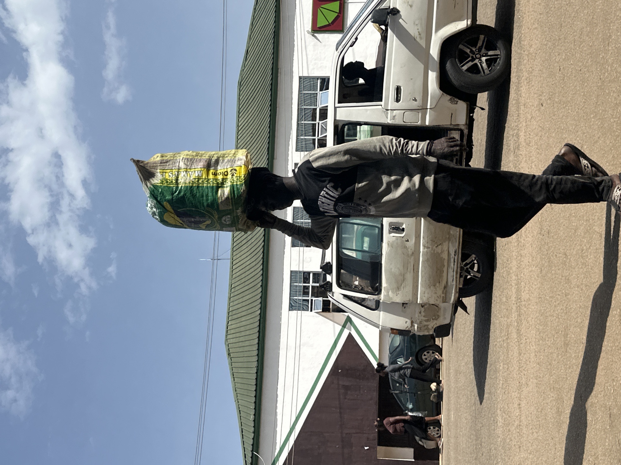 A young Black man walks down a dusty road in Bukuru, Nigeria, carrying a large sack of goods on his head. He is wearing a gray sweatshirt with black sleeves and a graphic design on the front, black pants, and sandals. The sack is green and yellow with some writing in a foreign language. The man is walking in front of a white van with its door open. A Black man is visible in the driver's seat of the van. Behind the van, there are two other vehicles and a building with a green roof. The sky is blue with some clouds. The sun is shining, casting a long shadow from the man onto the road. The scene appears to be a street market or commercial area.