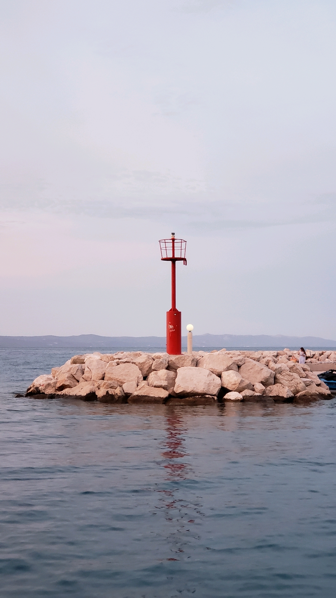 A vertical, low-angle, medium shot captures a red lighthouse standing on a rocky breakwater in the waters of Općina Tučepi, Croatia. The lighthouse is tall and thin, topped with a red cage-like structure. A small, round lamp is positioned below the cage. The breakwater is made of rough, light-colored stones, with a few larger rocks visible at the edge. The water is calm and dark blue, reflecting the lighthouse and the sky. In the background, a hazy line of hills or mountains can be seen across the sea. On the right side of the breakwater, near the edge, a person in a white dress is standing, facing away from the camera, and looking out at the water. Another person, partially obscured by a boat, can be seen further to the right. The sky is overcast and pale, with hints of pink and purple near the horizon, suggesting it may be dawn or dusk.