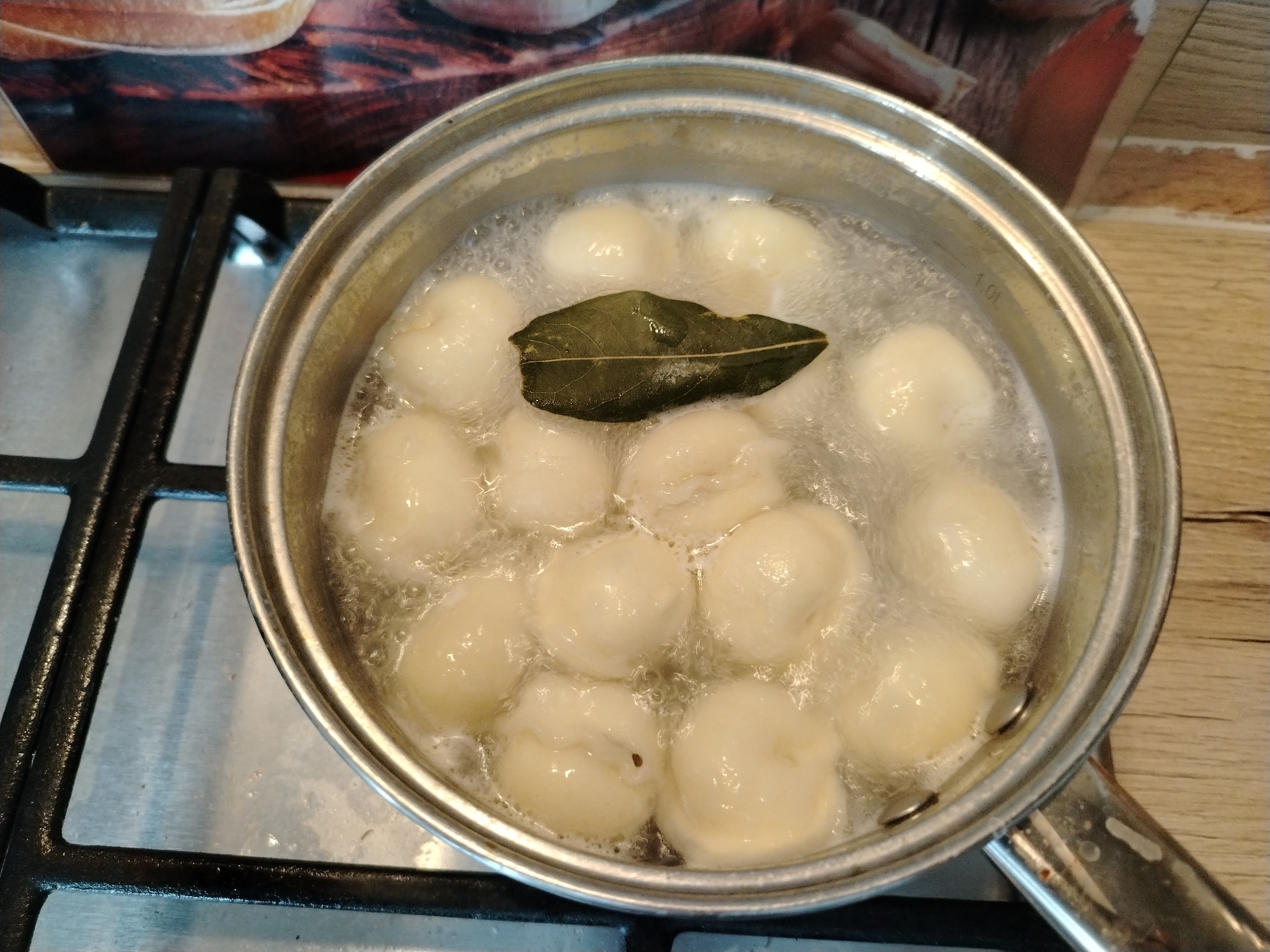 A top-down, close-up shot shows a stainless steel pot on a gas stovetop. The pot is filled with boiling water, and several round dumplings are cooking inside. A single bay leaf floats on the surface of the water. The stovetop is black metal, and the background includes a tiled backsplash with a reddish-brown and beige pattern. The scene appears to be set in a kitchen.