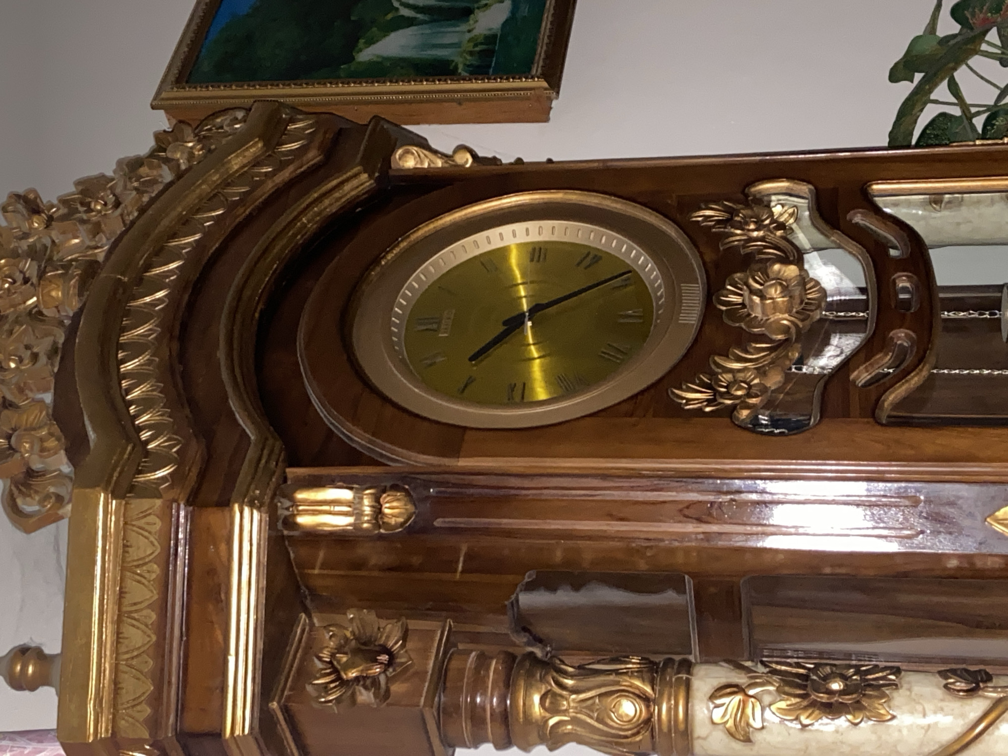 A close-up shot focuses on the upper portion of a tall, ornate wooden grandfather clock. The clock face, a golden color with Roman numerals and a thin black minute hand, is prominently displayed. Decorative gold embellishments, including floral motifs and carved patterns, adorn the clock's wooden casing. To the right of the clock, a framed painting depicting a waterfall and greenery is partially visible. The background is a plain white wall. The scene is indoors, with a hint of green foliage from a plant visible in the lower right corner. There are no people or discernible actions taking place. The lighting is bright, creating reflections on the polished surfaces of the clock. The image does not provide any information that would allow for the determination of its location in Margorejo, Indonesia.