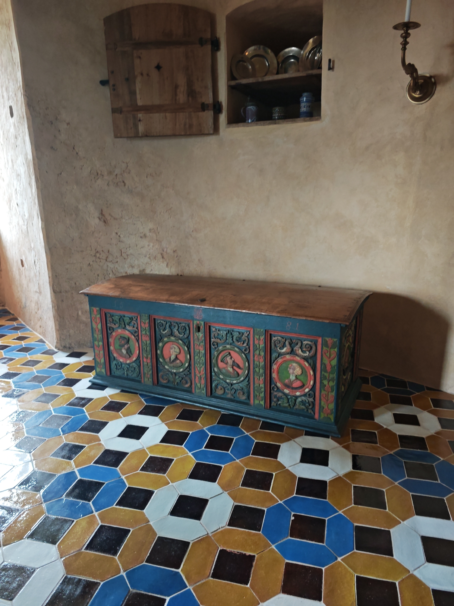The image depicts a room with historical architectural elements and furnishings, likely located in Bauska Castle, Bauska, Latvia. The scene is dominated by a large, intricately carved and painted wooden chest positioned on a floor tiled with a geometric pattern of blue, yellow, brown, white, and black octagonal and square tiles. The chest, painted predominantly in teal and red with green accents, features circular medallions with painted portraits on its front panels. Above the chest, a wall with a rough, textured finish features a small, recessed shelf displaying a collection of plates and two ceramic jars. To the left of the shelf, a wooden shutter with horizontal planks is mounted on the wall. A brass wall sconce with a candle holder is visible on the right side of the wall. The lighting suggests an indoor setting with natural light entering from the left.