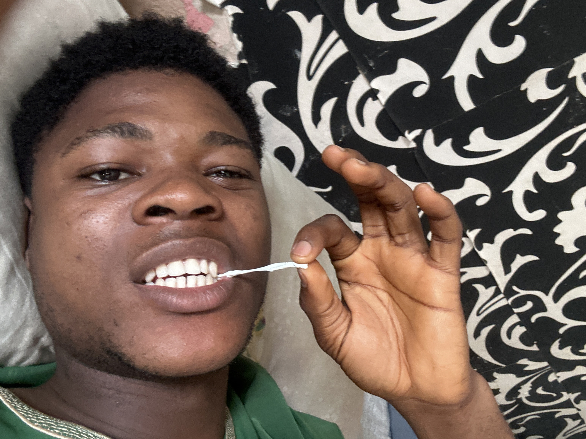 The media file is a close-up, selfie-style shot of a young Black person lying down, with a patterned fabric in the background. The subject is looking directly at the camera with an open mouth, revealing their teeth. They are holding a white, string-like object, possibly floss, between their fingers and inserting it into their mouth. The lighting suggests it could be daytime, indoors, given the absence of harsh shadows. The expression appears neutral, focused on the task at hand. The setting seems casual and domestic, indicated by the soft bedding and the patterned textile, which could be part of a couch or bedspread. The person is wearing a green garment. The location is identified as Ihiagwa, Nigeria. There is no visible text in the image.