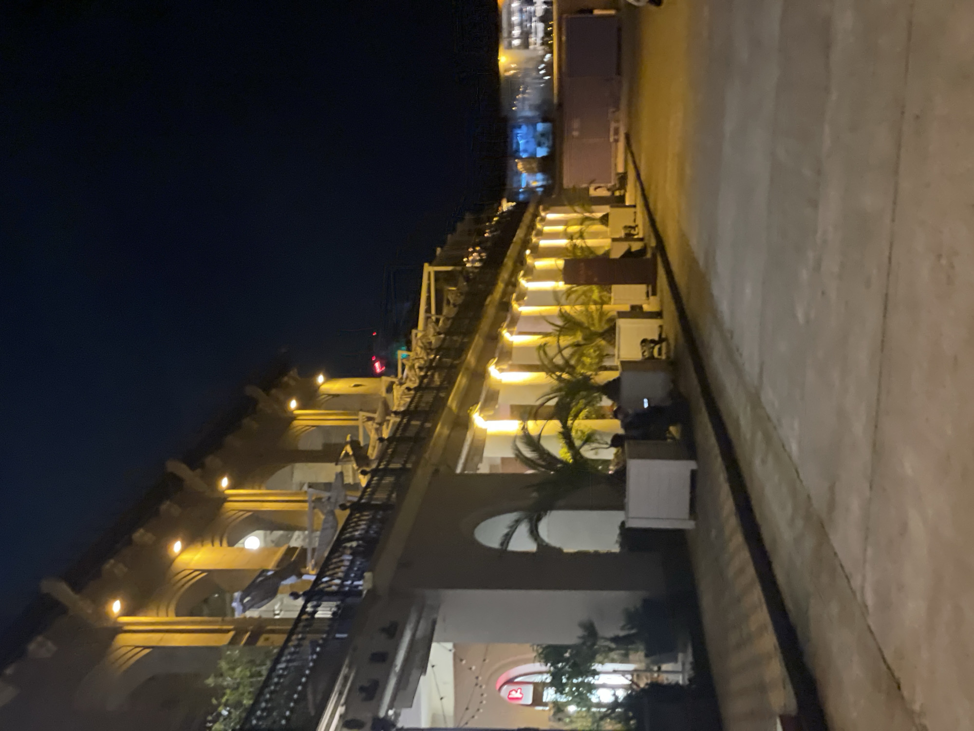 The image captures an outdoor scene at night in Sochi, Russia. The architecture features arches, lights, and balconies, possibly part of a restaurant or commercial building. Palm trees are planted in containers, adding greenery to the scene. A pedestrian walkway or sidewalk runs alongside the building. People are present, some sitting at tables, suggesting outdoor dining. The sky is dark, typical for a night scene. In the background, there are distant lights and structures, adding depth to the view.