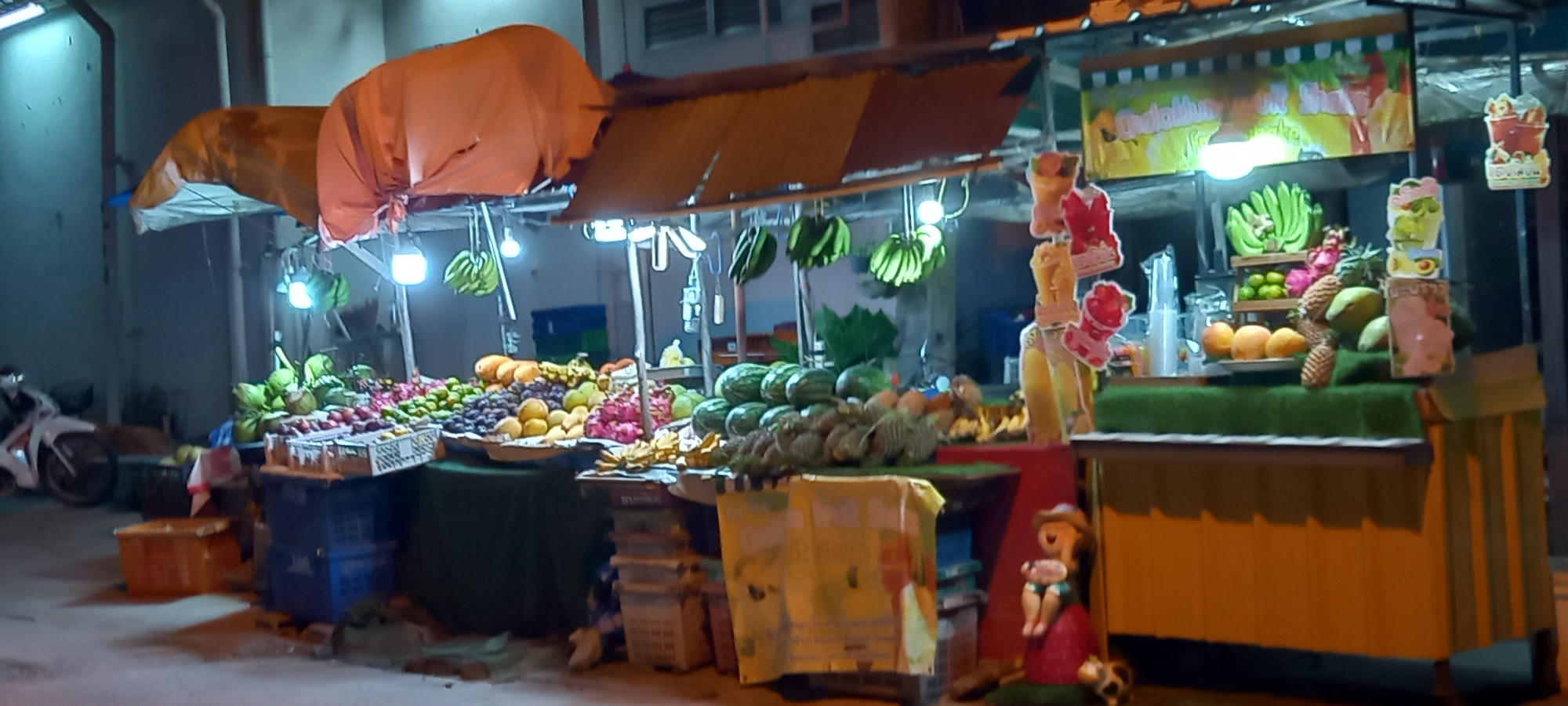 This nocturnal image depicts a vibrant fruit and juice stall set up along a street in Baan Chaloklum, Thailand. The stall is illuminated by several bright hanging lights, casting a glow over the diverse array of fresh tropical fruits on display.
The main subject is the fruit stall, characterized by orange and brown awnings that shelter the produce. Various fruits are meticulously arranged on tables and in crates. On the left, green coconuts, dragon fruit, grapes, and mangoes are visible, along with what appear to be mangosteen and rambutan. Towards the center and right, large watermelons are stacked, alongside pineapples, durian, bananas hanging from hooks, and limes. The general activity is the sale of fresh fruit and prepared fruit shakes.
Notable details include the time of day, which is clearly night due to the dark surroundings and the reliance on artificial lighting. The stall features a dedicated section for making fruit shakes, indicated by blenders and cups, and decorated with colorful cut-outs of fruit smoothies. A yellow banner above this section prominently displays the text "Ohatakliua Fruit Shake". On the far right, a small decorative figurine of a child or gnome sits near the base of the stall. On the far left, partially visible, is a white motorcycle parked on the street. The scene evokes the lively atmosphere of a Southeast Asian night market.