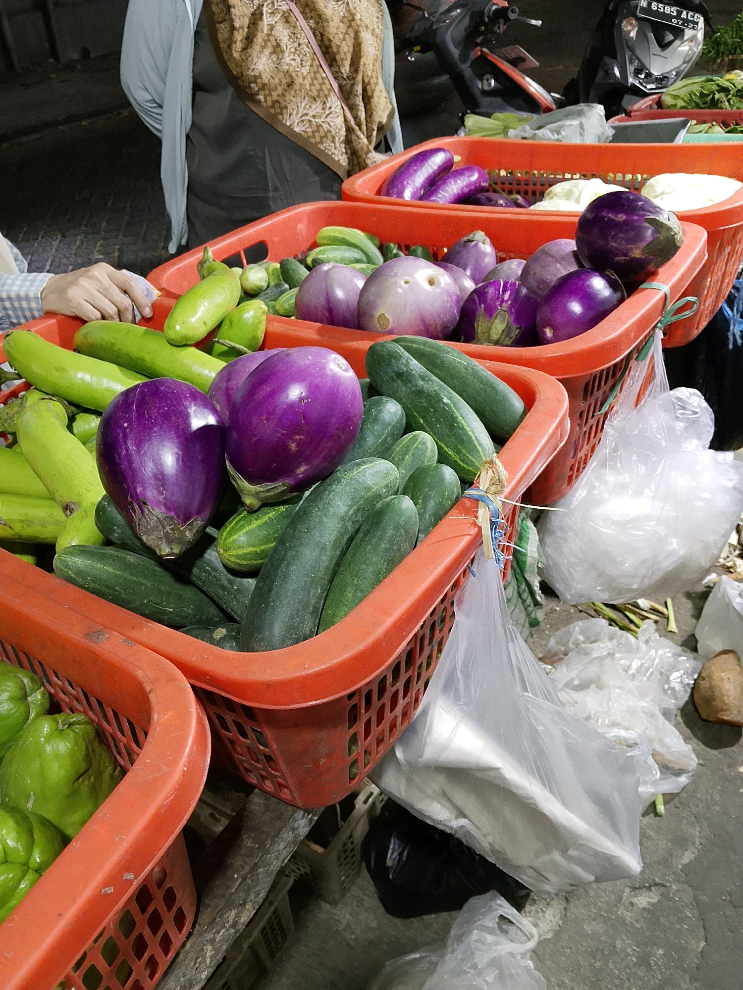 This is a close-up, eye-level outdoor photograph taken at a street market in Malang, Indonesia. The image focuses on several bright orange plastic baskets filled with fresh produce. In the foreground, a basket is packed with vibrant purple eggplants and dark green cucumbers. To the right, another basket contains more eggplants, some appearing lighter purple and others a deeper violet. Behind these, a larger basket holds long, light green gourds or beans, mixed with some lighter green, rounder vegetables that could be chayote or a similar squash.
A person's arm, wearing a light blue and white checkered long-sleeved shirt, is visible reaching into the basket on the left, holding a purple item. Further in the background, a woman is partially visible, wearing a brown patterned headscarf and a light blue top. A motorcycle with a black body and a white license plate is parked behind the baskets. The ground appears to be concrete or asphalt, with some debris and plastic bags scattered around. The lighting suggests it is daytime, possibly late afternoon or early evening, with some shadows cast on the ground. The overall scene depicts a typical produce stall at a bustling Indonesian market.