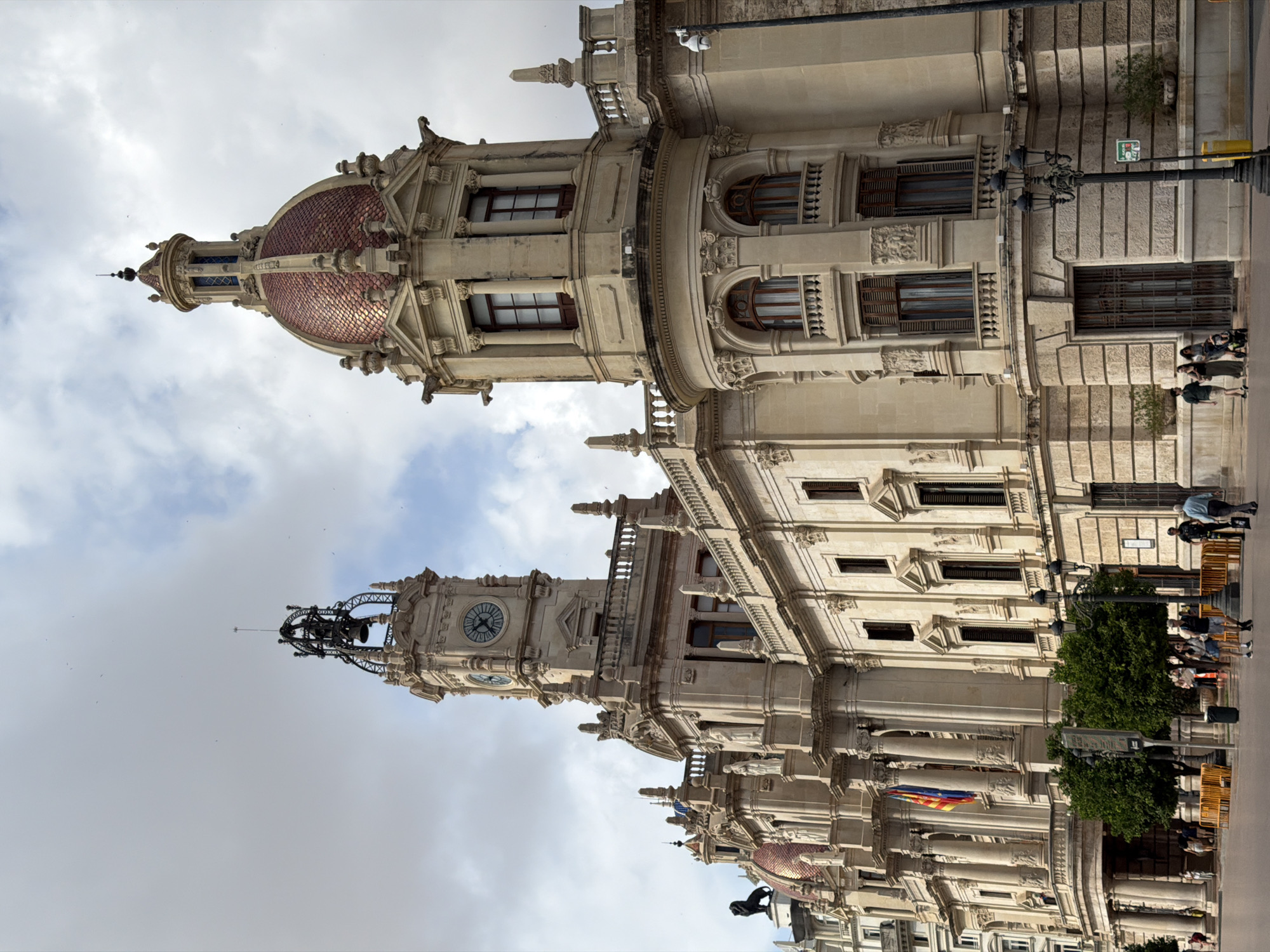 This image depicts the City Hall (Ayuntamiento) of Valencia, Spain, under a partially cloudy sky. The grand, multi-story building features a light-colored stone facade with intricate architectural details. On the left, a prominent clock tower rises, topped with an ornate black metal bell structure and a slender spire. A circular clock face is visible on the tower. To the right, a large dome covered in reddish, possibly copper, tiles dominates, surmounted by a smaller cupola and a finial.
The building's facade is adorned with numerous windows, some featuring small balustraded balconies and dark wooden shutters. Columns, pilasters, and decorative carvings are integrated throughout. A portico with large columns and a balcony is visible in the mid-left, from which the national flag of Spain and the flag of the Valencian Community are displayed. A dark lion statue is perched on the building's roofline to the left of the clock tower. Gargoyle-like embellishments are visible near the base of the main dome. A white security camera is mounted on a black pole to the far right, near the building.
The foreground consists of a paved public plaza or street. Several small, green-leafed trees are planted along the front of the building. Black, ornate street lamps are positioned throughout the foreground and midground. Yellow protective barriers line parts of the plaza. Multiple pedestrians are present, walking individually or in small groups. Some individuals are near the trees, others closer to the building's entrance. One person is pushing a stroller. A person in a uniform, possibly a security officer or police, stands near the right side of the frame.
