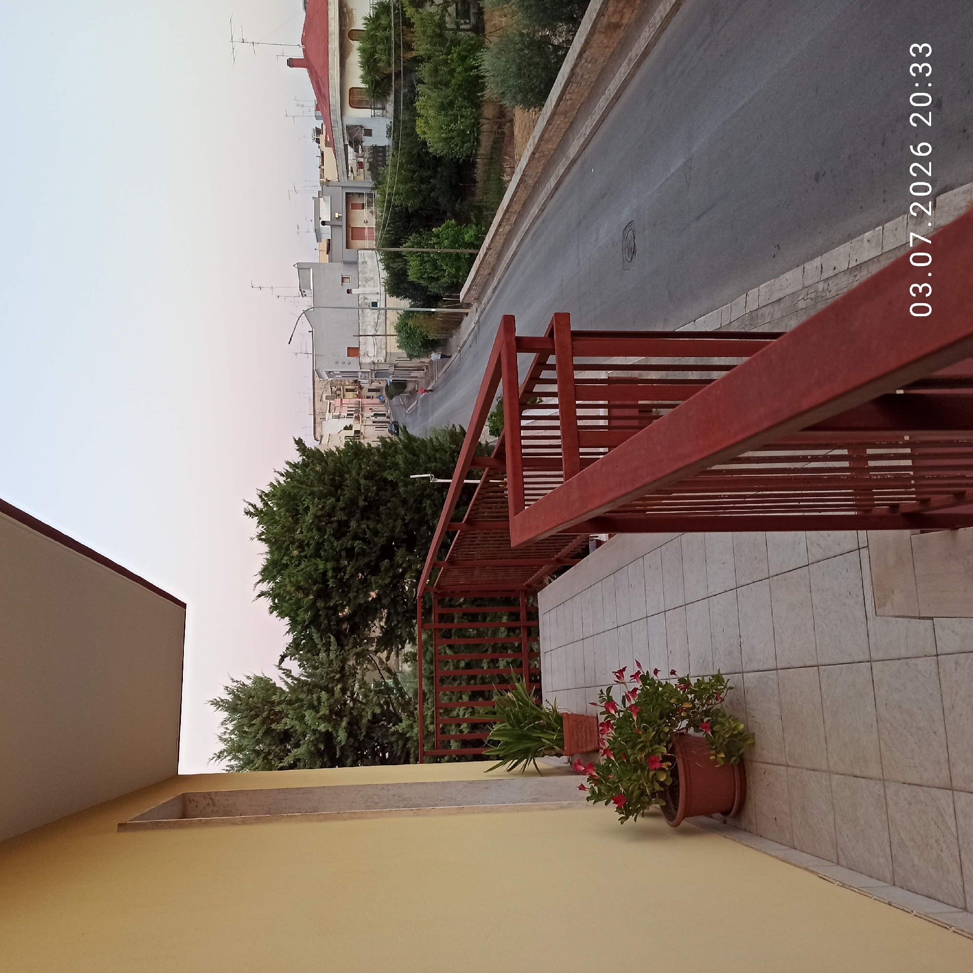 The image shows a view from a balcony in Bitonto, Italy, overlooking a street lined with buildings and trees. A red railing frames the foreground. On the balcony, there are two potted plants: one with green foliage and another with red flowers. The street is paved and appears to be a residential area, with a few cars parked or driving. The sky is a pale pinkish-blue, suggesting either sunrise or sunset. Buildings of various heights and architectural styles are visible on both sides of the street. The timestamp "03.07.2026 20:33" is overlaid on the image.