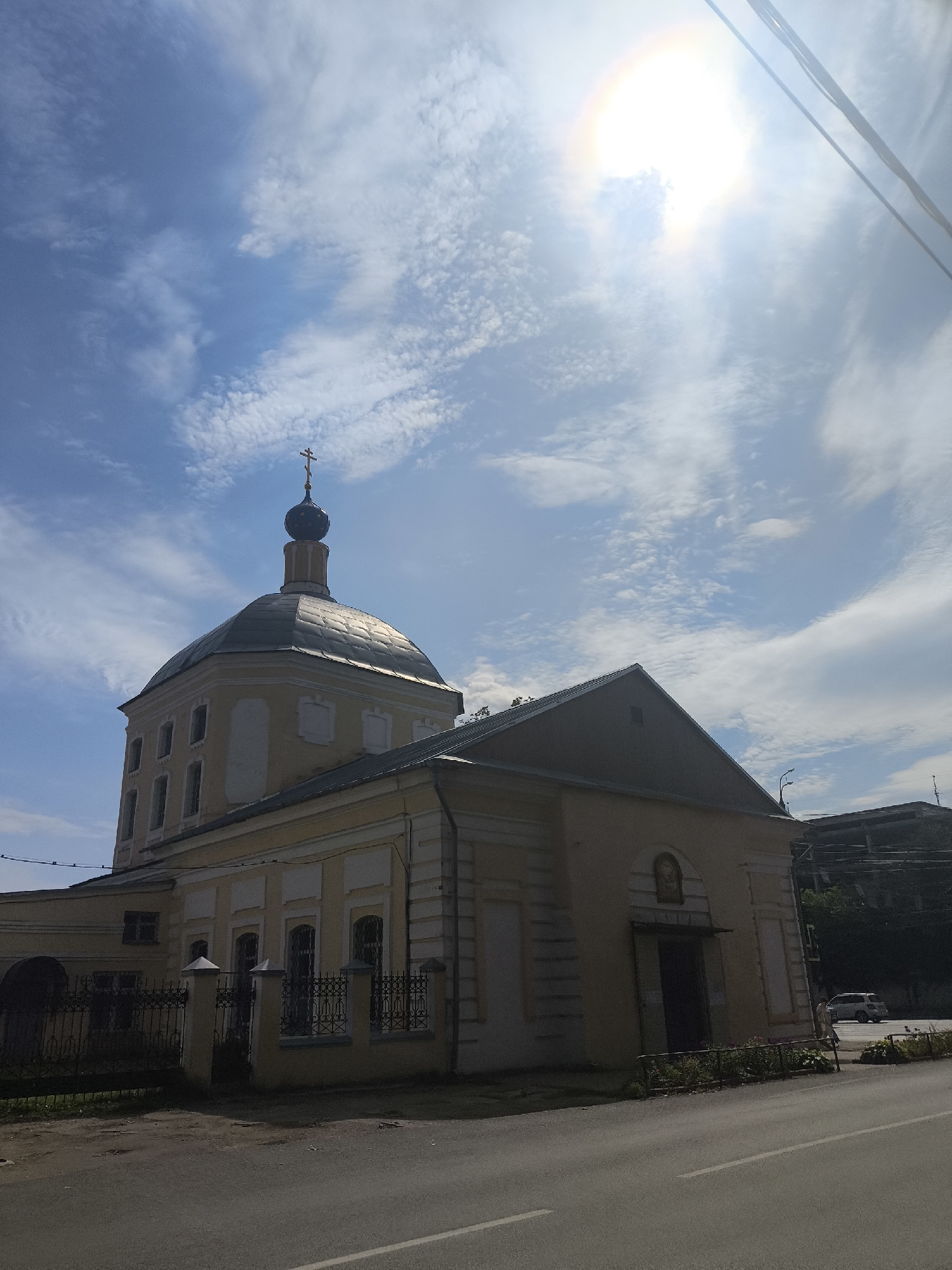 An exterior, eye-level, long shot captures a yellow, stone-built church with a prominent dark dome topped by a cross. The church is situated next to a road with a white line dividing the lanes. The sky is a bright blue with scattered white clouds, and the sun is intensely bright, partially obscured by wires in the upper right corner, creating a halo effect. A black wrought-iron fence runs along the front of the church. Beyond the church, a few trees and buildings are visible. A white car is parked on the side of the road, and a person can be seen walking near it. The scene is illuminated by natural daylight, suggesting a daytime setting.