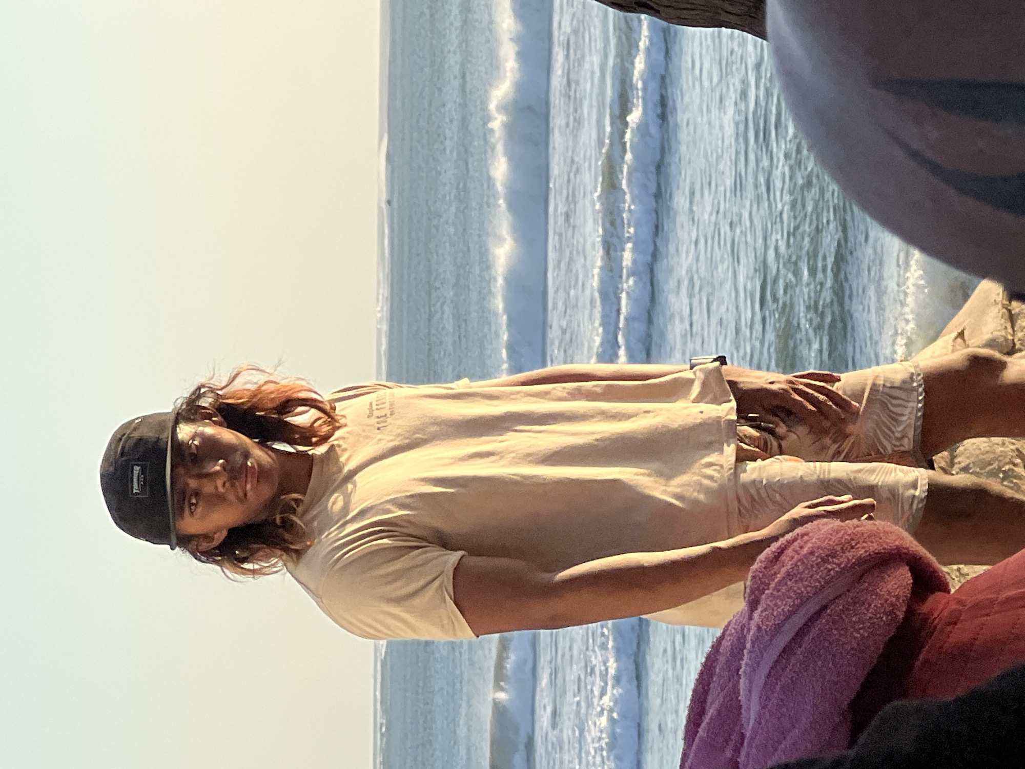 The image shows a young man wearing a black baseball cap and a tan t-shirt with shorts standing on a rocky shoreline. The ocean is behind him with waves breaking. The sky is clear and light, suggesting it is either sunrise or sunset. The man has long, reddish-brown hair and is looking directly at the camera. In the foreground, a portion of a pink towel or blanket is visible, along with another person's shoulder and head. The overall setting appears to be a beach in Kuta, Indonesia, with the characteristic golden light of the Balinese coast.