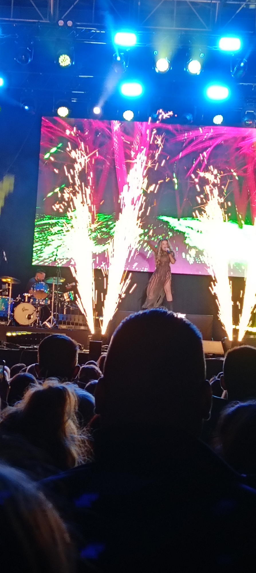 The image shows a live musical performance taking place in Cerdanyola Del Vallès, Spain. A female singer is the main subject, standing on a stage and singing into a microphone. There are pyrotechnic effects shooting up on either side of her, suggesting an energetic and visually dynamic performance. In the background, a large screen displays abstract patterns in pink and green. To the left of the singer, a drummer can be seen playing a drum set. A crowd of people are watching the performance, with the backs of their heads and shoulders visible in the foreground, indicating the perspective of someone in the audience. Above the stage, stage lighting is visible.
