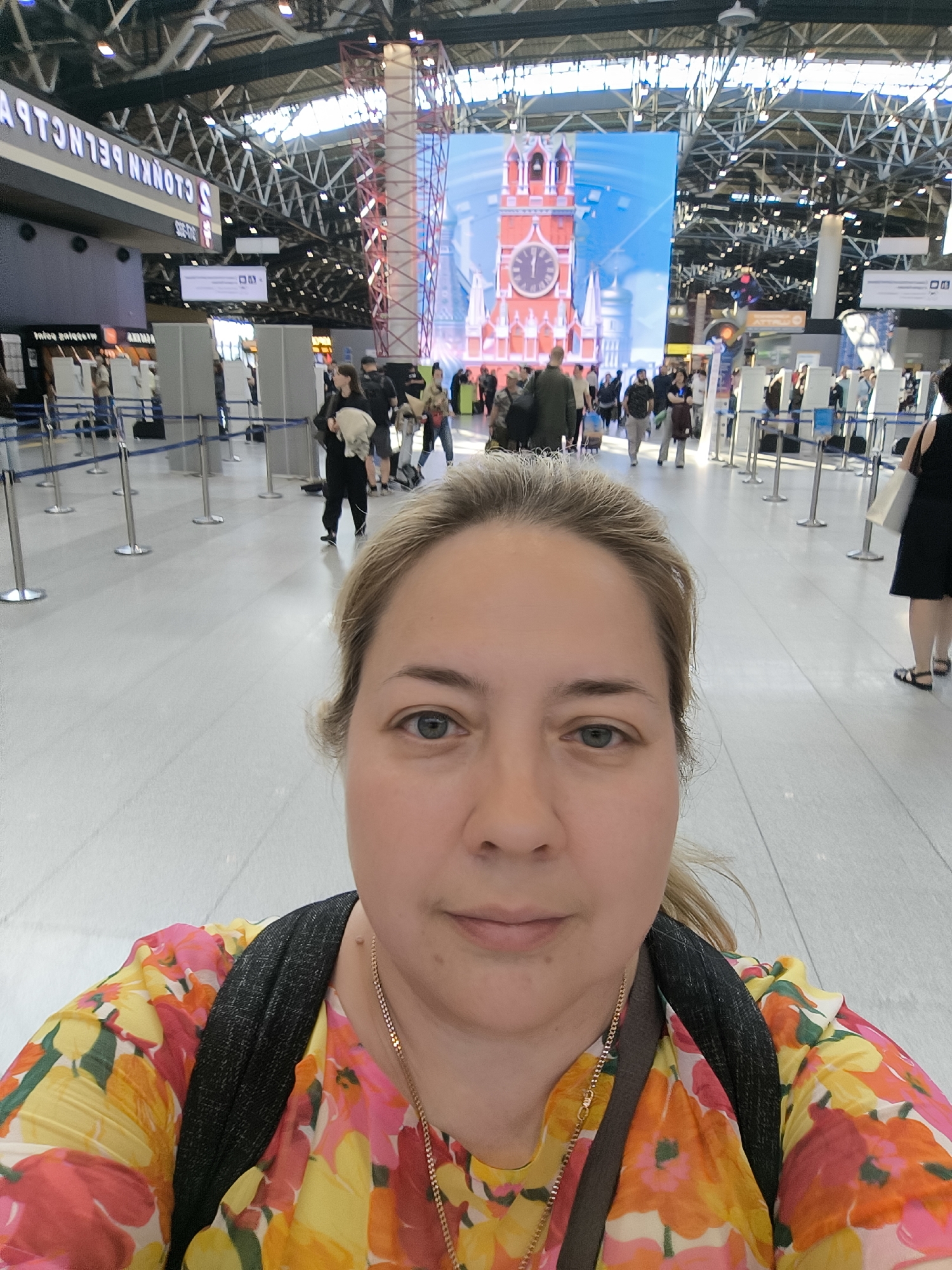 A female individual with light-colored hair and blue eyes is captured in a selfie in the foreground, facing the camera directly. She wears a brightly colored top adorned with yellow, orange, pink, and red floral patterns, along with a dark shoulder strap from a backpack and a thin gold chain necklace.
The background reveals the interior of a busy airport terminal, identified as Sheremetyevo International Airport in Khimki, Russia, by mirrored Russian text "С ШЕРЕМЕТЬЕВОМ" (With Sheremetyevo) on an overhead sign. Numerous passengers are visible in various states of movement, some walking, others potentially queuing near white self-service kiosks. These areas are delineated by silver metal stanchions connected by blue retractable belts.
A prominent large digital display screen in the mid-background features an illustration of a red clock tower, strongly resembling the Spasskaya Tower of the Moscow Kremlin, set against a bright blue sky. The terminal architecture includes a high ceiling with exposed metal trusses and beams, illuminated by overhead lighting fixtures. The floor is uniformly light-colored and tiled. Several individuals carry luggage, and one person in the mid-left appears to be carrying a light-colored, possibly wrapped, item.