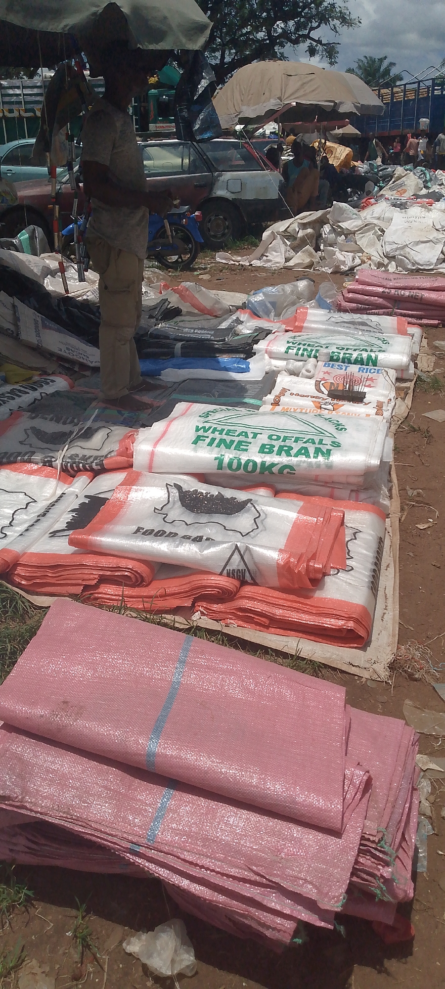 A market scene in Tor-Donga, Nigeria is depicted with a focus on stacked woven sacks. In the foreground, a pile of pink woven sacks with a blue stripe is visible, partially obscuring other sacks. Behind these, a layer of white and orange woven sacks is arranged, some bearing green lettering that reads "WHEAT OFFALS FINE BRAN 100KG" and other indistinct text. Further back, additional stacks of similar sacks are visible. A person, partially obscured by the sacks and a large umbrella, stands to the left. In the background, several cars, including a white station wagon and a blue truck, are present, along with what appears to be more market stalls and trees. The ground is a mixture of dirt and scattered debris. The overall impression is one of a bustling open-air market.