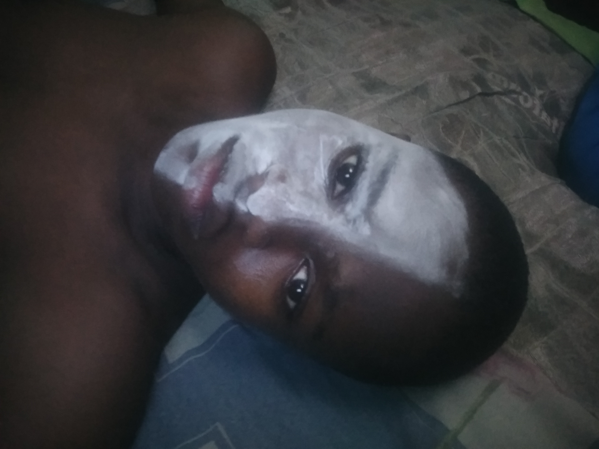 The image captures a close-up, angled shot of a person, likely a child or young adult with dark skin, lying down indoors. Their head rests on what appears to be a blue-patterned pillow or bedding. A prominent feature is the white substance applied unevenly across a large portion of their face, covering the forehead, part of the nose, and cheeks, resembling a facial mask or face paint. Their dark eyes are open and look directly towards the viewer. The person's bare shoulder and a portion of their upper torso are visible, suggesting they are shirtless. The background shows textured, possibly grey or brown, bedding or a mattress, with faint patterns. Hints of blue and green fabric items are also visible in the periphery. The lighting is dim, suggesting a low-light environment, possibly evening or night, or a dimly lit room. The person's expression is neutral. There is no visible text in the image.