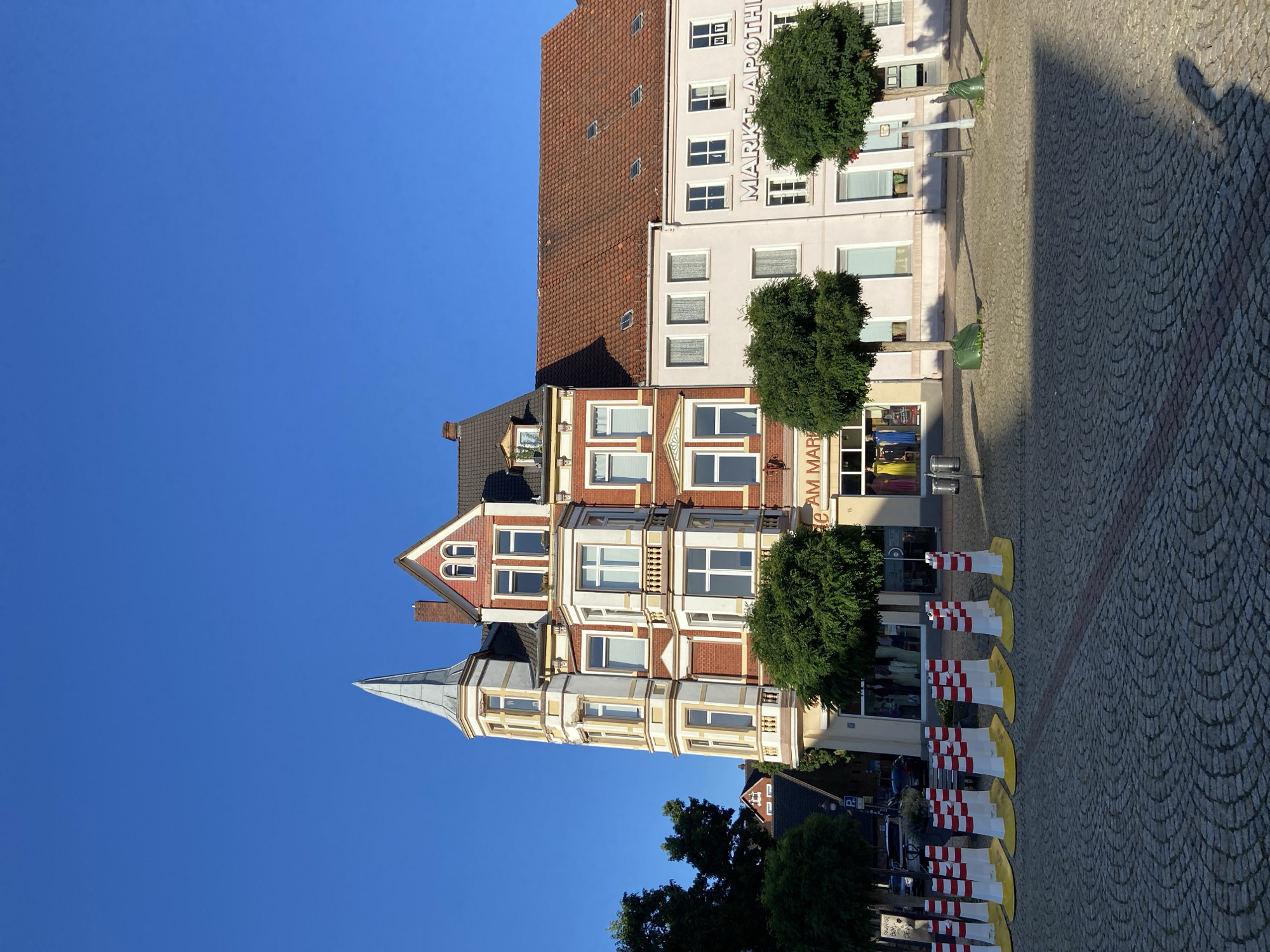 A clear blue sky is above a street in a European city. In the center of the frame is a multi-story building with a red brick facade. The building has a bay window and a tall, pointed turret. To the right of this building is a lighter-colored, three-story building with a red tile roof. This building has windows and a sign that reads "MARKT-APOTHEKE." In front of the buildings is a cobblestone square. A row of yellow and white striped traffic cones lines the edge of the square, and trees are planted in front of the buildings. Cars are visible on the street behind the cones.