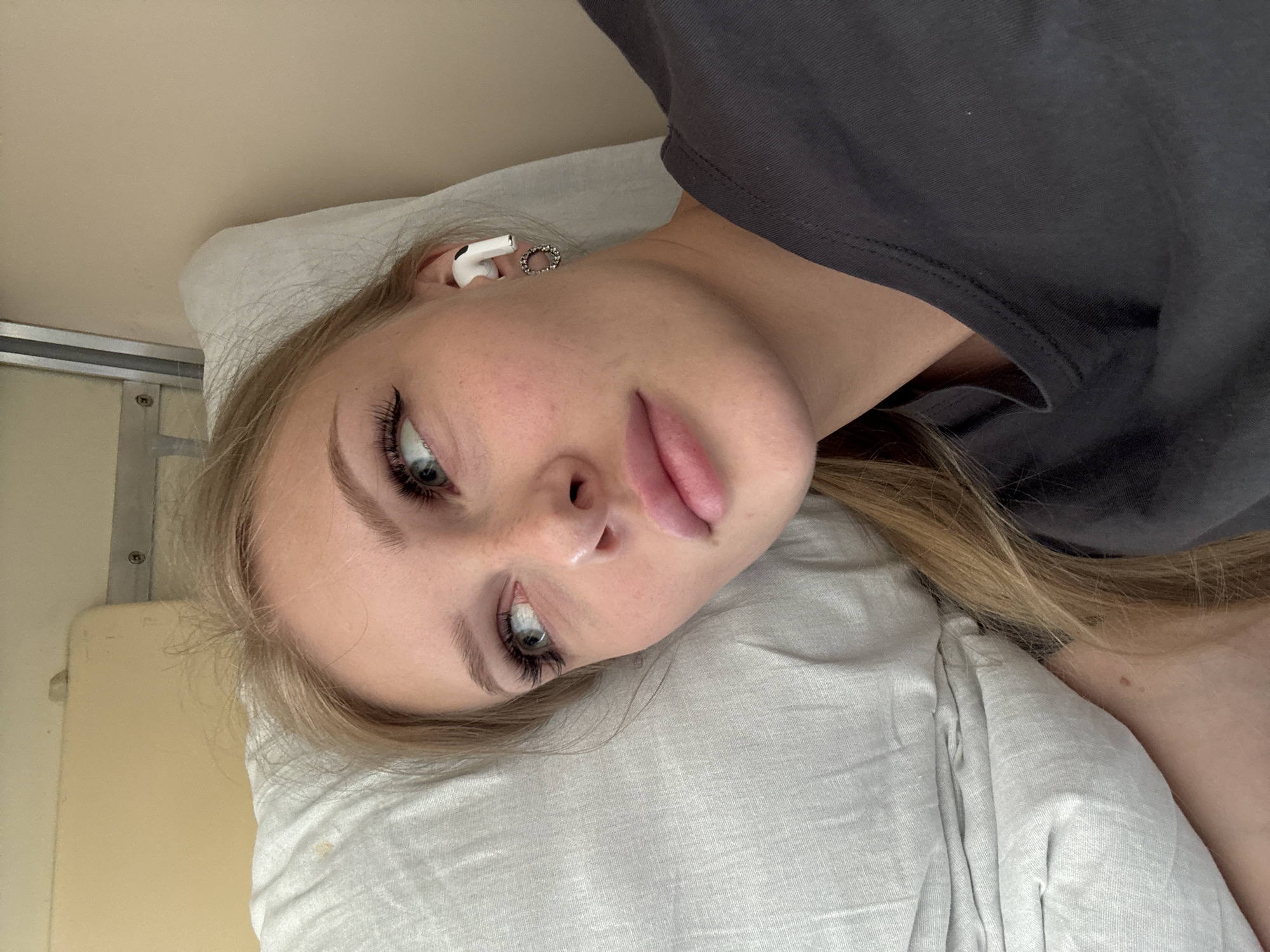 A young woman with blonde hair, wearing a dark t-shirt and an earbud, is lying on a pillow on a train. She is looking to her left, with her eyes partially closed and her lips slightly parted. The background appears to be a train interior, with a beige wall and a metal railing visible.