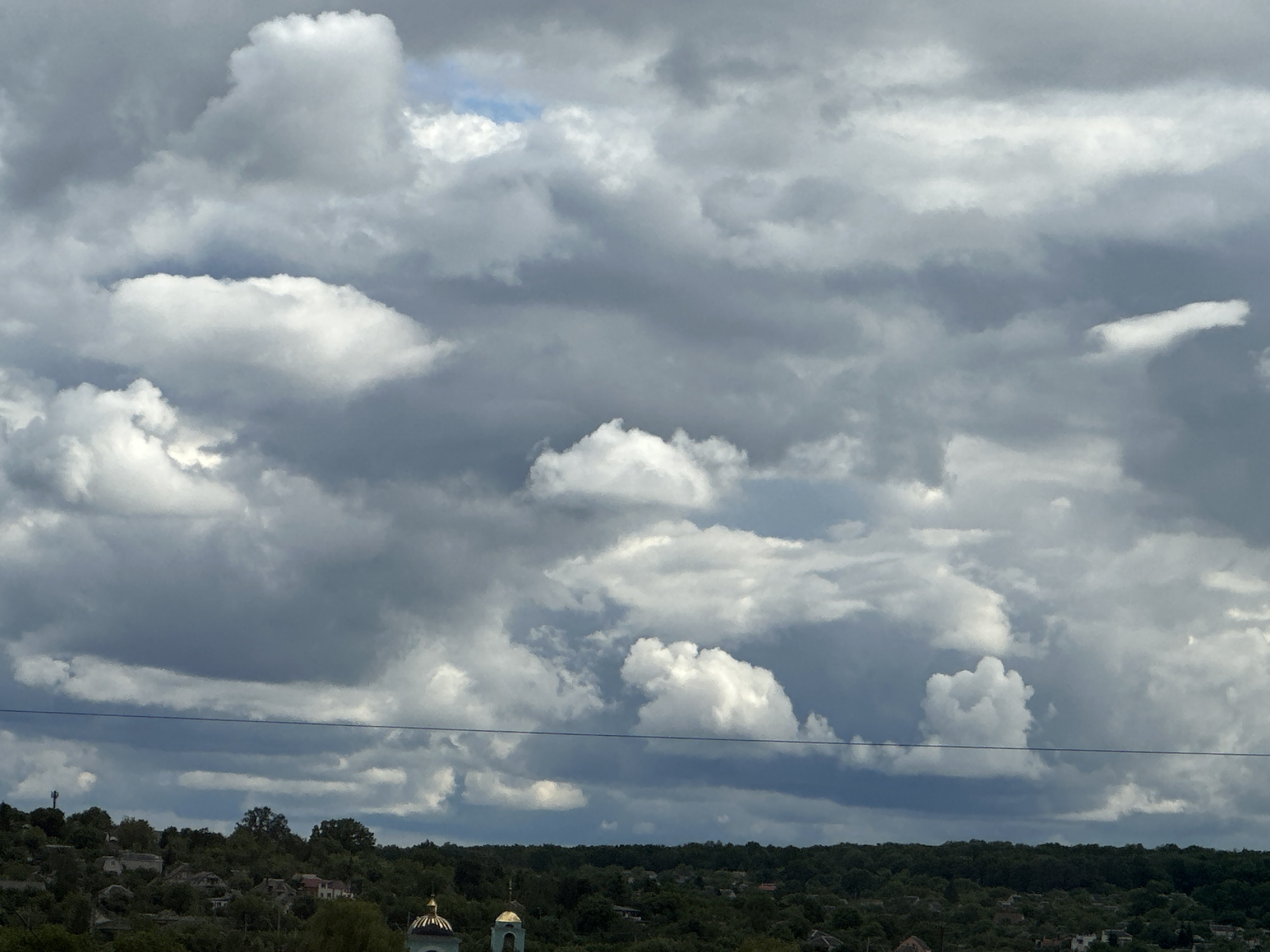 The image shows a cloudy sky over a landscape in an unknown city. In the foreground, there is a line of trees and rooftops of buildings. Two golden domes of a church are visible. The sky is filled with grey and white clouds, with a small patch of blue sky visible.