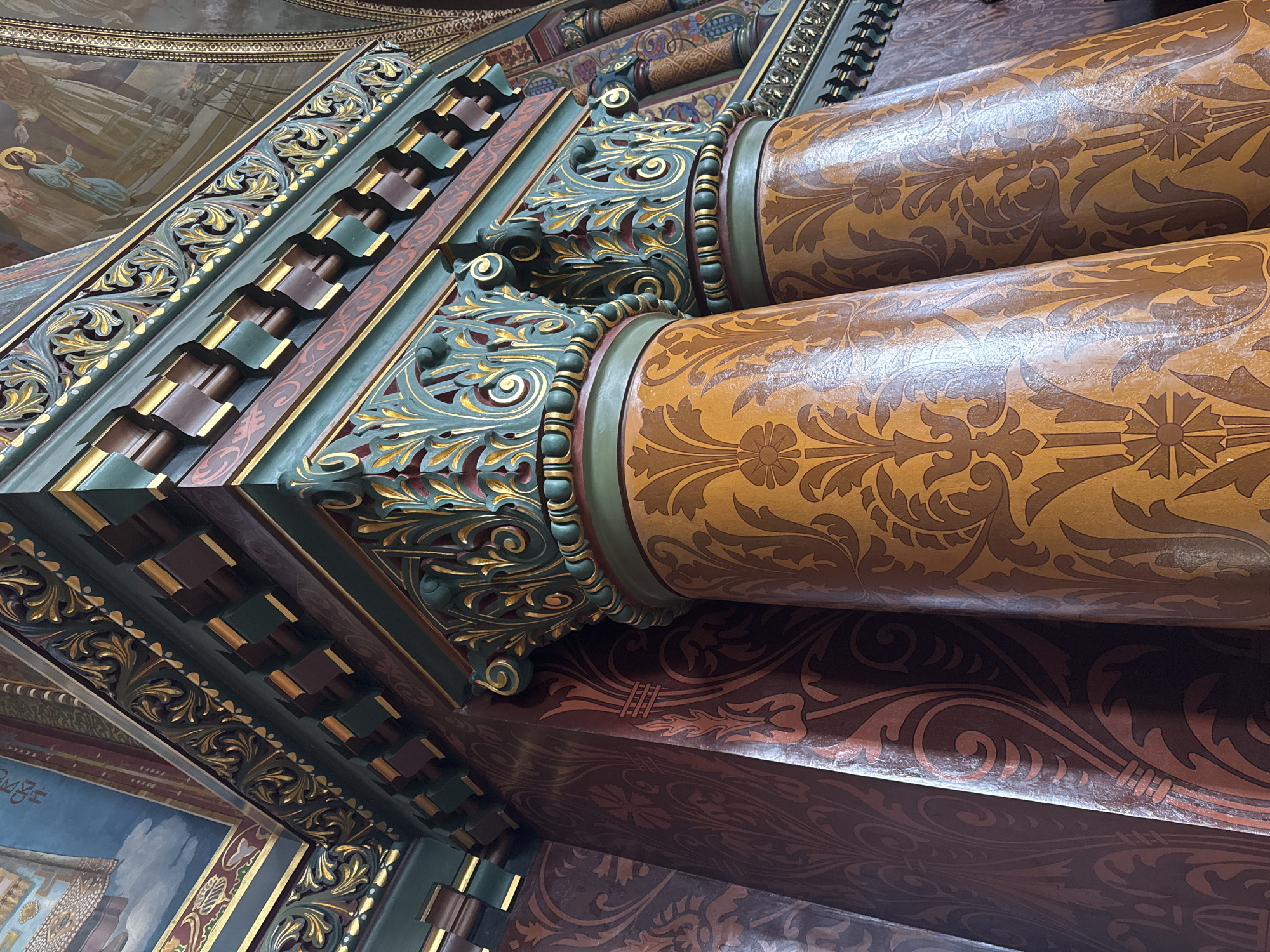 The image captures a section of elaborate interior architecture, likely within a historical building. The primary elements are two cylindrical columns adorned with a floral pattern in shades of brown and tan. These columns are capped with intricate capitals featuring a mix of green, gold, and brown ornamentation, including scrolling leaf designs and beaded bands. Above the columns, a portion of a ceiling structure is visible, featuring a stepped design with brown, green, and gold accents. In the upper left corner, a segment of a fresco painting can be discerned, showing figures in robes. The location is likely Saint Petersburg, Russia, given the style of the architecture and decoration, and it suggests an interior of a church or palace.