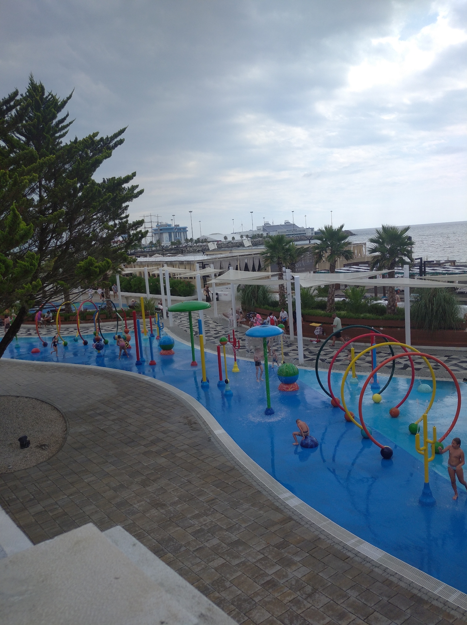 This image depicts a children's water play area on a cloudy day, likely at a resort or public park in Sochi, Russia. The water play area features a bright blue surface with various colorful spray features, including mushroom-shaped sprinklers and archways with hanging balls. Several children are visible, interacting with the water features. Some are running through the sprays, while others are sitting or standing in the shallow water. In the background, palm trees and a beach area with lounge chairs and umbrellas suggest a coastal location. Further back, a large building with a ship-like structure docked nearby, along with several tall poles, indicates a port or harbor. The overall scene is one of recreational activity on an overcast day, with the sea visible in the distance.