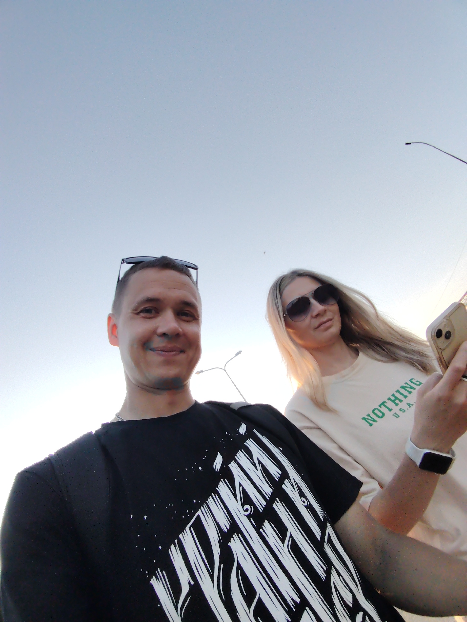 The image shows two individuals, likely in Chaikovsky, Russia based on prior context. A man with short hair and sunglasses atop his head is in the foreground, wearing a black t-shirt with a white geometric design. He is smiling. Behind him, a woman with long blonde hair and sunglasses is holding a smartphone. She wears a light-colored shirt with "NOTHING U.S.A." printed on it. A white smartwatch is on her left wrist.
The background is a clear sky, suggesting an outdoor location. Street lights are visible, and the overall lighting implies either early morning or late afternoon.