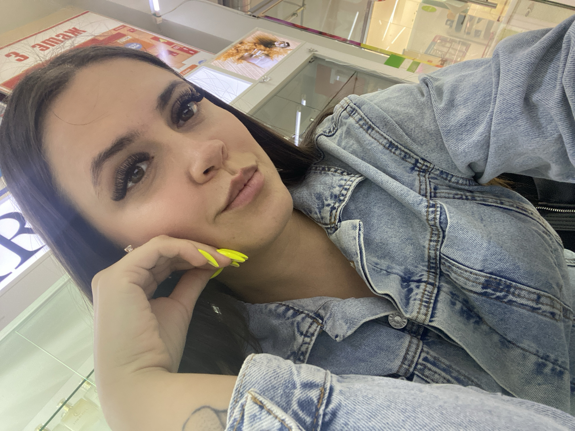 A woman is in a store in Chaikovsky, Russia, posing for a selfie. She has long dark hair, dark eyebrows, and long eyelashes. She wears a light blue denim jacket. Her fingernails are painted neon yellow. She rests her face on her left hand, where a ring is visible. Glass display cases are visible behind her, filled with merchandise. Text in Russian can be seen on a sign above the display cases.