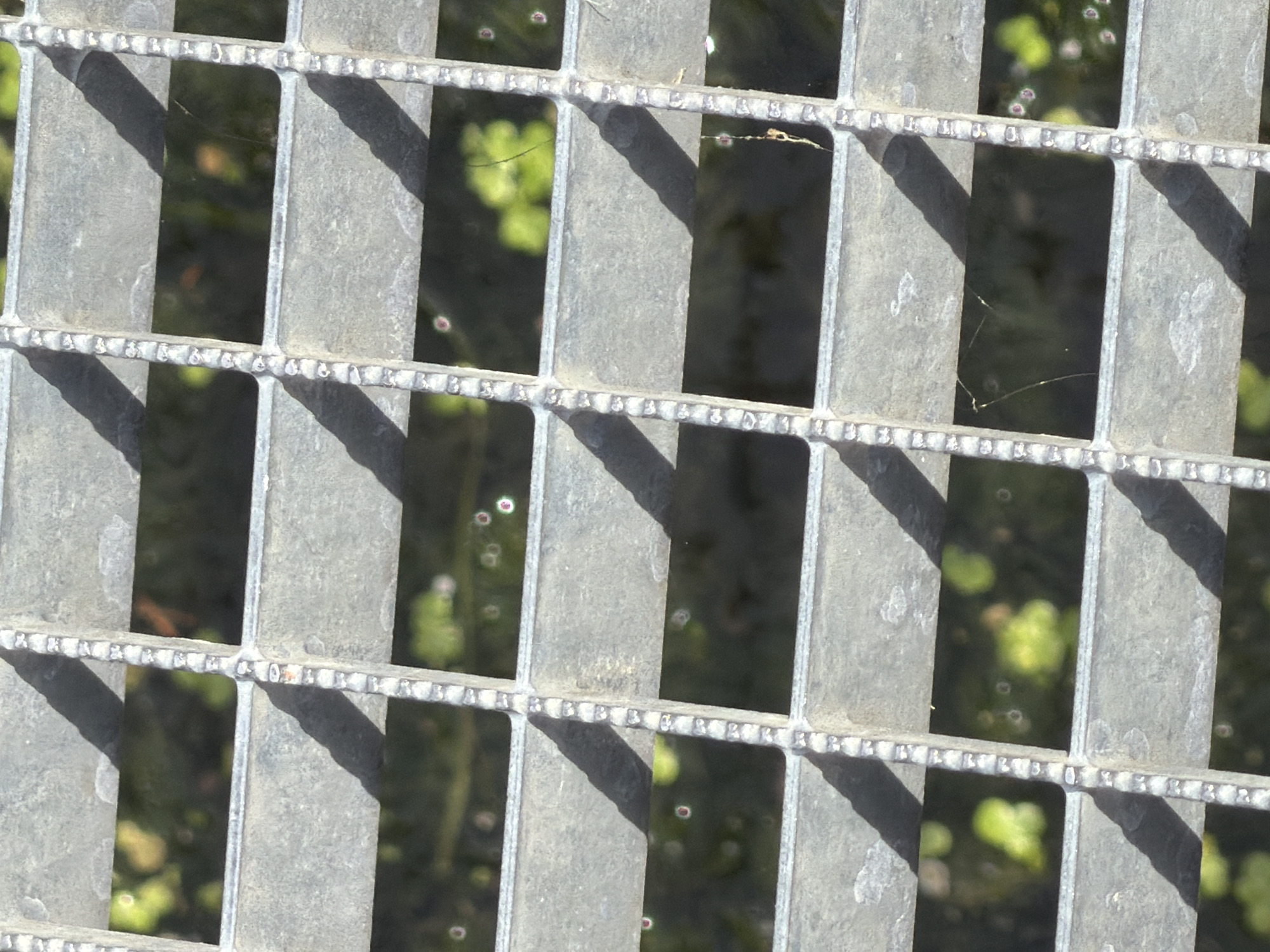 The image shows a close-up view of a metal grate, possibly a footbridge or a ventilation cover, in Joinville-le-Pont, France. The grate consists of horizontal and vertical metal bars, with the metal appearing weathered and somewhat dirty. Behind the grate, out-of-focus foliage and vegetation can be seen, suggesting an outdoor environment. Sunlight casts distinct shadows across the grate from the bars, indicating a bright, sunny day. There are no people or animals visible in the image, nor are there any identifiable structures or specific landmarks of Joinville-le-Pont. The overall scene is focused on the texture and pattern of the metal grate and the blurred natural elements behind it.