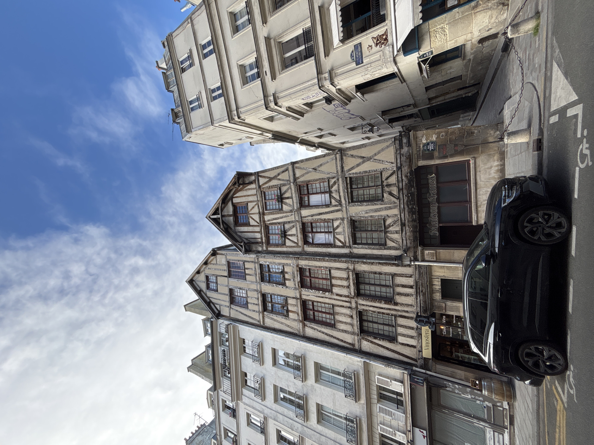 The image captures a street scene in Paris, France, characterized by historic architecture. In the foreground, a black SUV is parked on the street, partially obscuring a sidewalk and a chain barrier. The street features yellow and white markings, including a directional arrow and a wheelchair symbol. The buildings lining the street showcase a mix of architectural styles. On the left, a pale, multi-story building has numerous windows with decorative iron balconies. Adjacent to it are two narrow, older buildings with prominent timber framing and darker window frames. The building on the right features a light-colored facade with multiple windows and balconies, and at its base, awnings and signage for businesses. One sign reads "Maison Faust". Above the buildings, a blue sky with scattered white clouds is visible. The overall impression is of a charming, historic Parisian street.