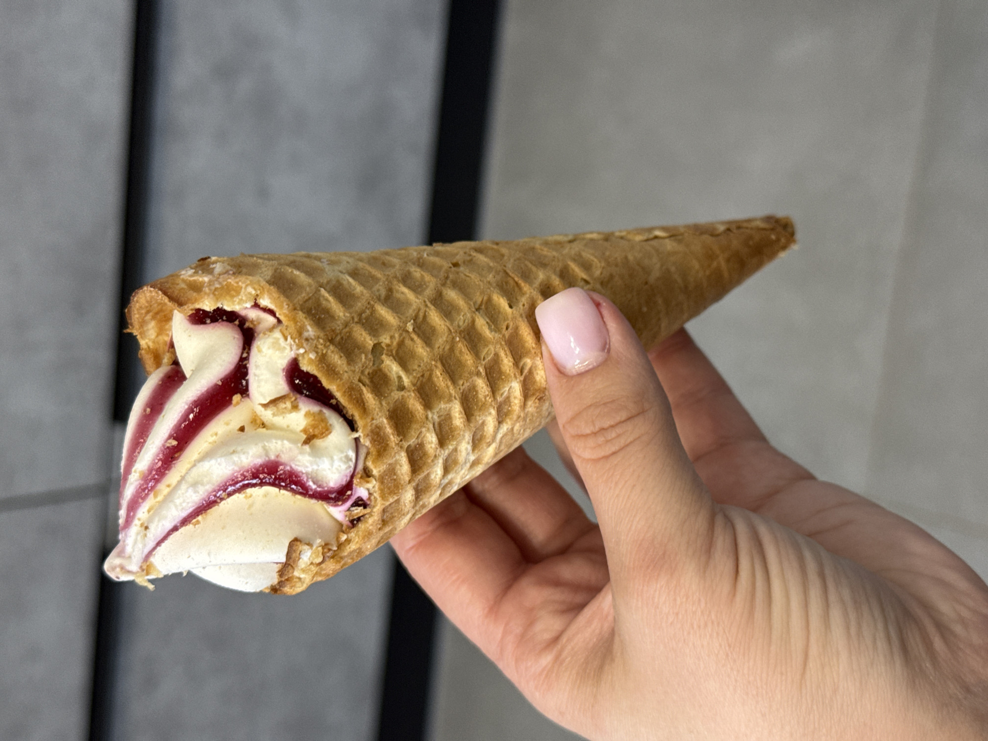 A person's hand, with light skin and a light pink nail polish, holds a waffle cone filled with white ice cream swirled with a red berry sauce. The cone is golden brown and textured. The background is a blurry gray wall with horizontal lines. The scene is indoors. The location is not discernible from the image.