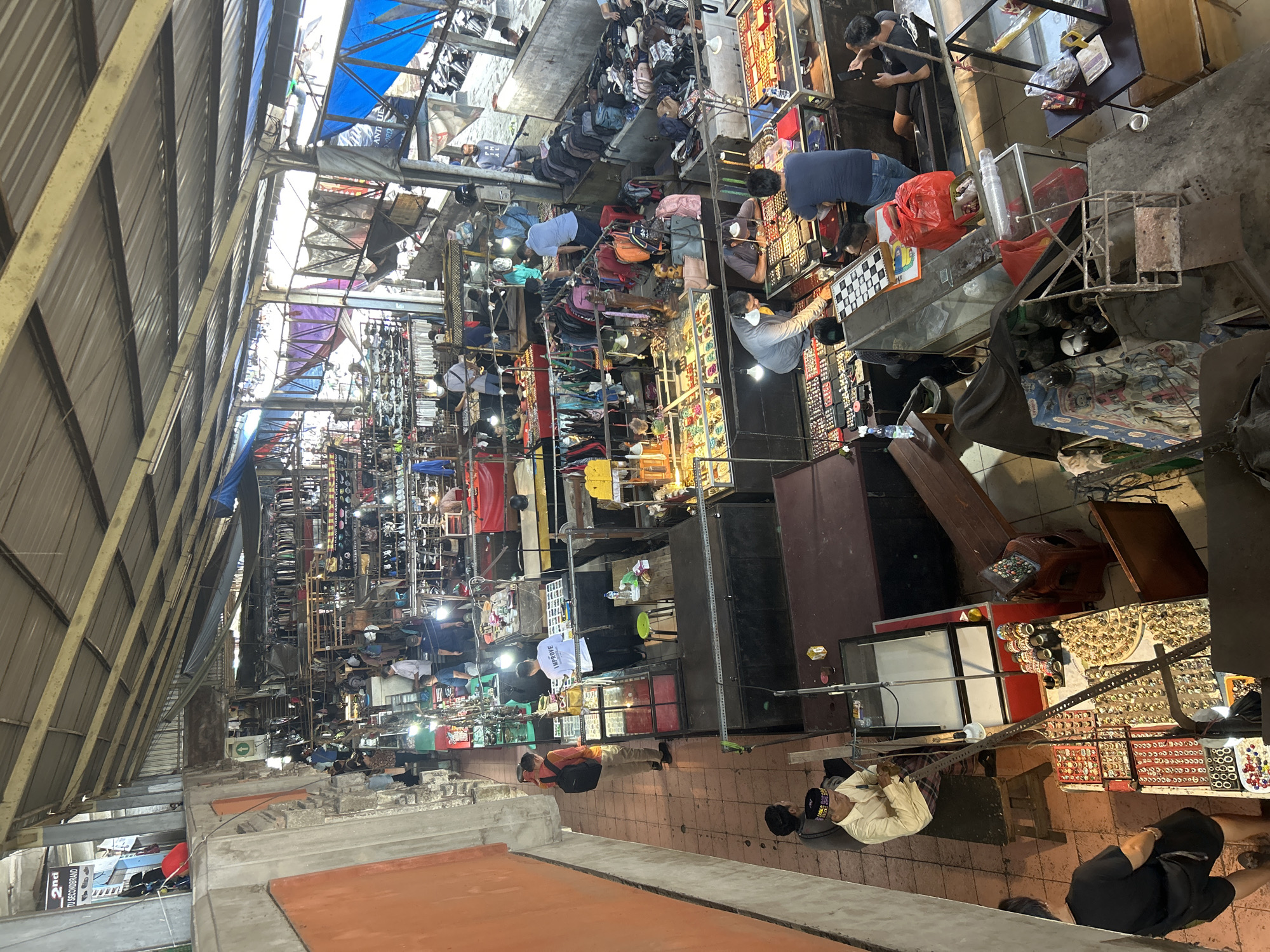 This image depicts a busy indoor market in Denpasar, Indonesia, likely a flea market or a second-hand goods market, given the variety of items on display. The market appears to be structured with numerous stalls arranged under a corrugated metal roof supported by metal beams. The stalls are packed with merchandise, including clothing on racks, bags, jewelry displayed in glass cases, and various other assorted items.
In the foreground, several individuals are seen browsing or tending to stalls. One man in a white shirt with "IMPROVE" printed on the back is looking at jewelry displayed in a glass case. To his left, another man wearing khaki pants and an orange shirt with a backpack is also looking at jewelry. Further down the aisle, a group of men are gathered around a stall featuring a chessboard and various small items in cases.
The market is illuminated by overhead fluorescent lights, casting a bright, somewhat harsh light on the scene. The overall atmosphere is one of bustling commerce, with people actively engaged in shopping and selling. The architecture suggests a functional rather than decorative space, designed to maximize the number of stalls and their visibility. The abundance of goods and the density of people indicate a lively and active marketplace.