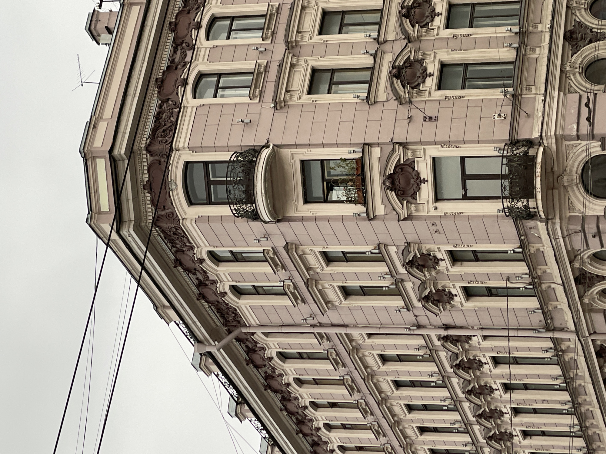 The image shows the exterior corner of a building in Saint Petersburg, Russia. The building has a light pink facade with ornate architectural details around the windows, including decorative moldings, carvings, and balconies with wrought-iron railings. The windows are rectangular with arched tops. A few electrical wires are visible against the gray sky in the background.