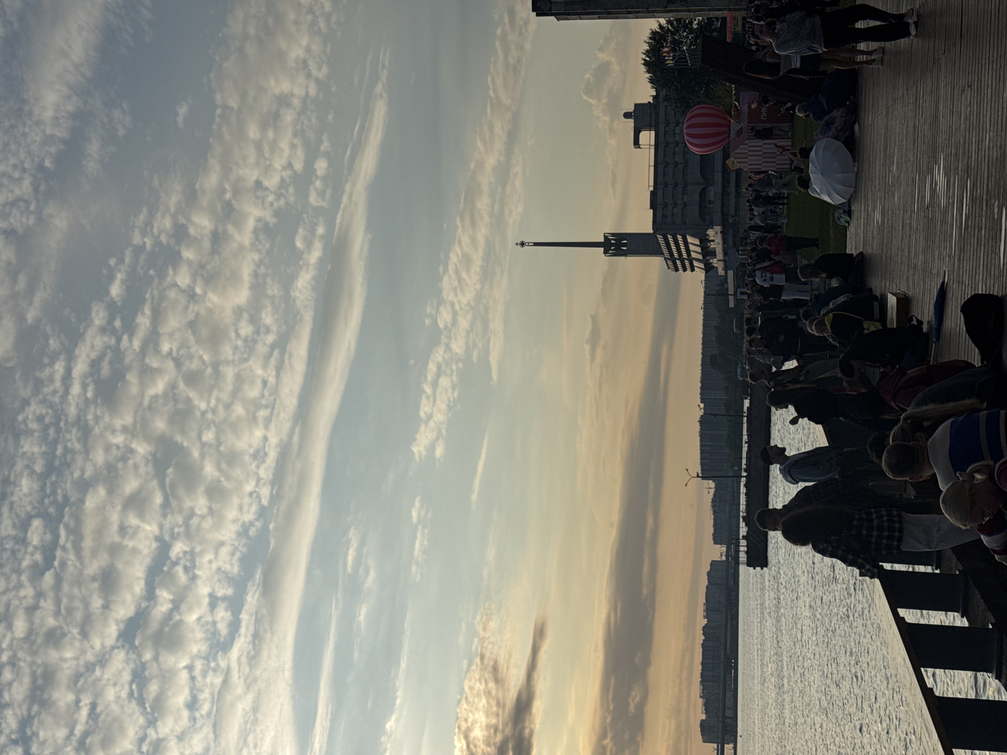 The image captures a sunset scene at the Sevkabel Port in Saint Petersburg, Russia. A body of water, likely the Gulf of Finland, occupies the left side of the frame, with a wooden pier and walkway forming the foreground. Numerous people are gathered on the pier and the grassy area behind it, some sitting, some standing, and some appearing to be in conversation.
Prominent structures in the background include a distinctive building with a tall, spire-like structure on top, and a large, striped hot air balloon. The sky is filled with scattered clouds, illuminated by the warm hues of the setting sun, creating a dramatic and atmospheric backdrop. The overall impression is one of a public gathering or event taking place during a pleasant evening.