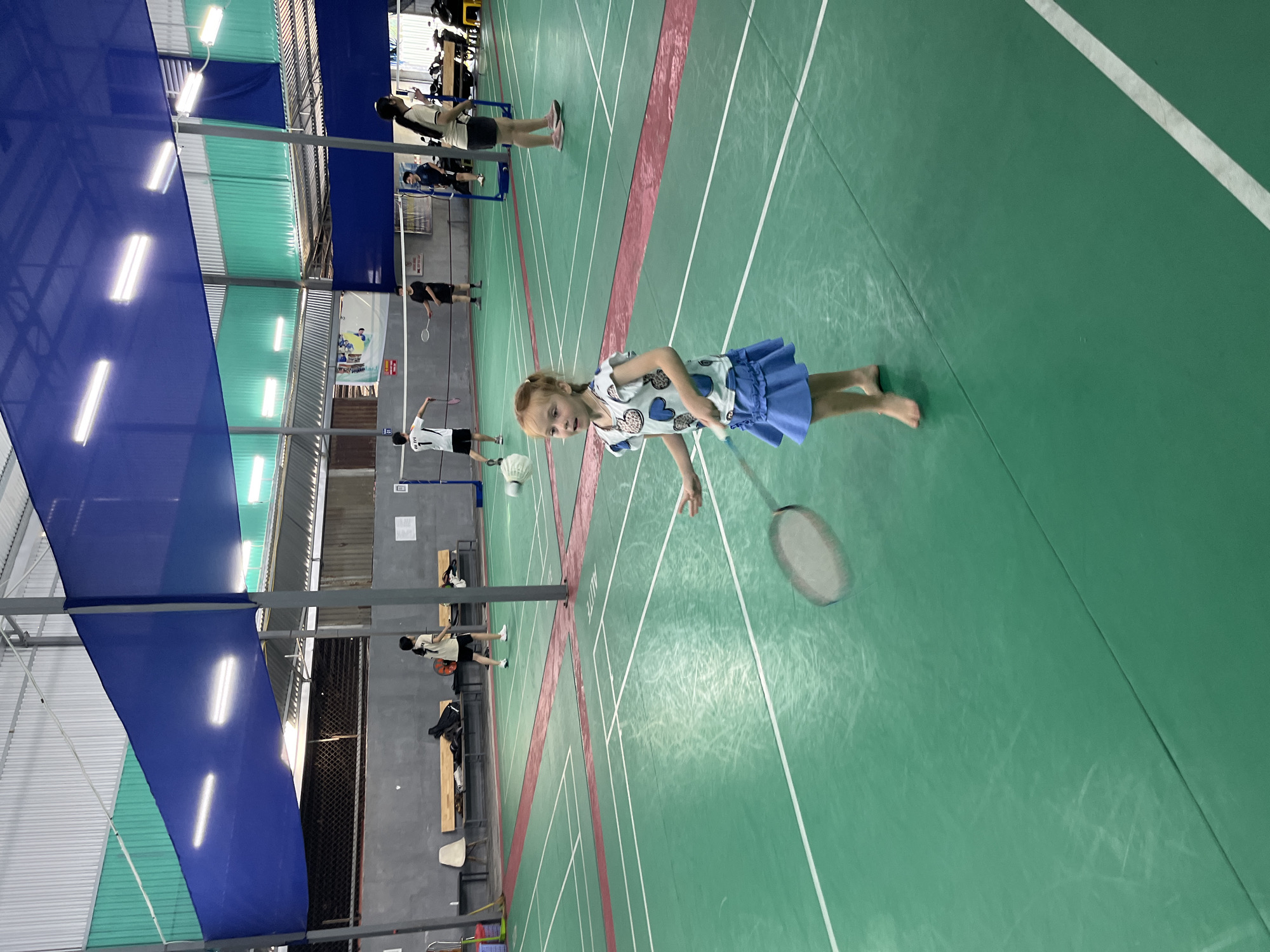 This image depicts an indoor badminton court in Bac Nha Trang Ward, Vietnam. The court is marked with green flooring and red boundaries. In the foreground, a young Caucasian girl with blonde hair is standing barefoot, wearing a white t-shirt with blue heart patterns and a blue skirt. She is holding a badminton racket, with the racket's head blurred due to motion, suggesting she is playing or about to play. In the background, several individuals, likely Vietnamese, are engaged in playing badminton on adjacent courts. The environment includes metal support structures, a blue net-like canopy overhead, and fluorescent lighting. Benches and bags are visible along the perimeter of the court.